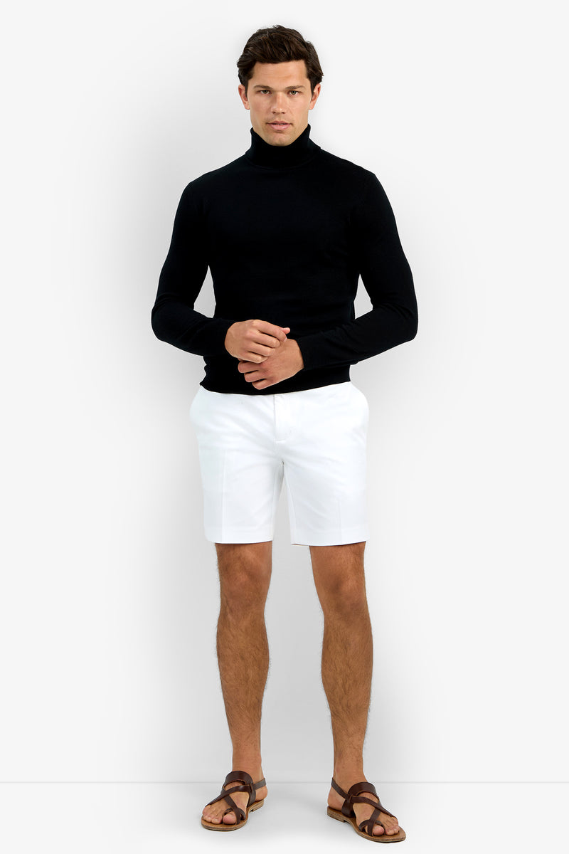 A person stands against a plain background wearing the Roy Long Sleeve Turtleneck in black, paired with white shorts and brown sandals. They have short brown hair and are looking forward with hands clasped together.