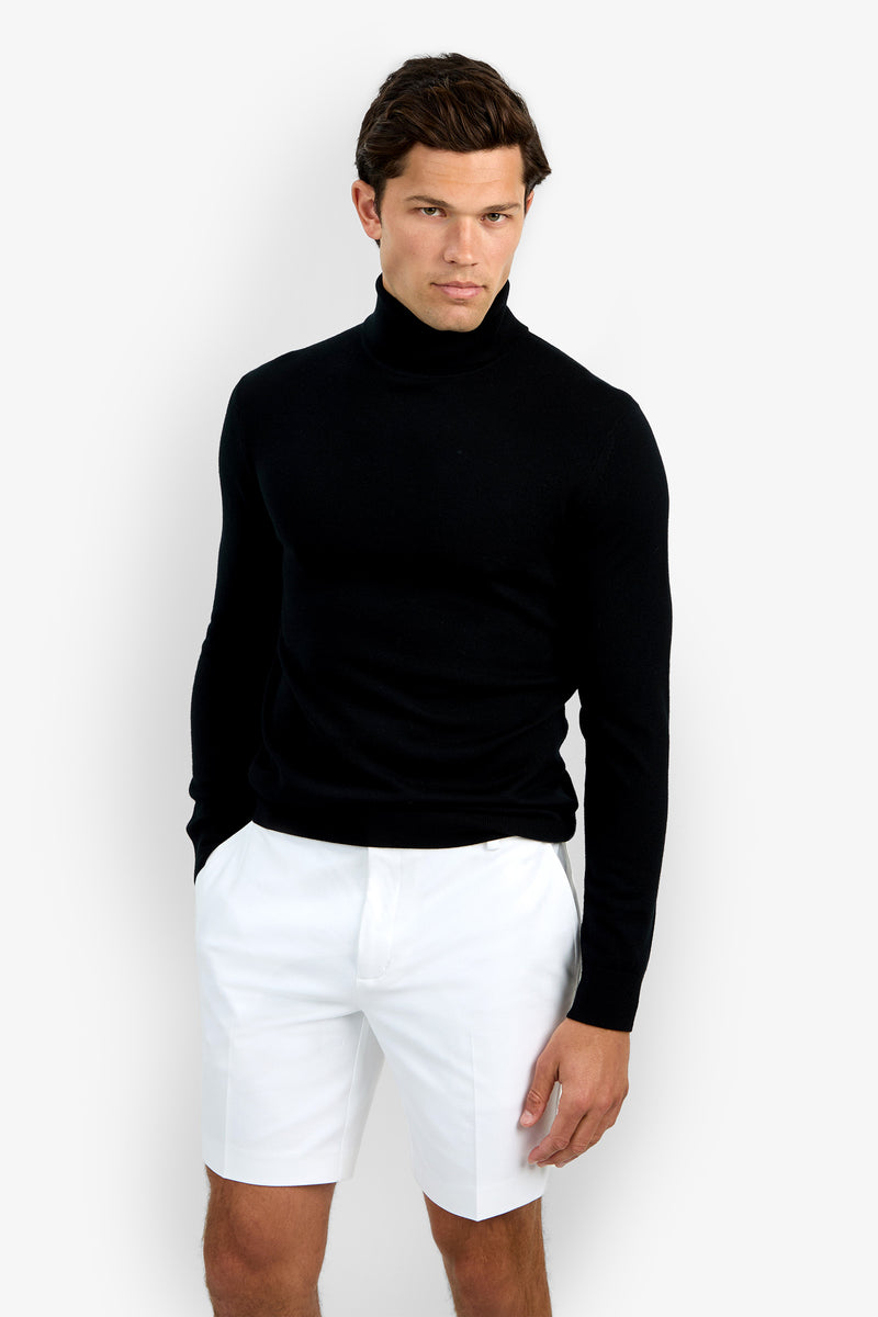 A man stands against a plain white background, wearing the Roy Long Sleeve Turtleneck in black and white knee-length shorts, with one hand in his pocket and a neutral expression.