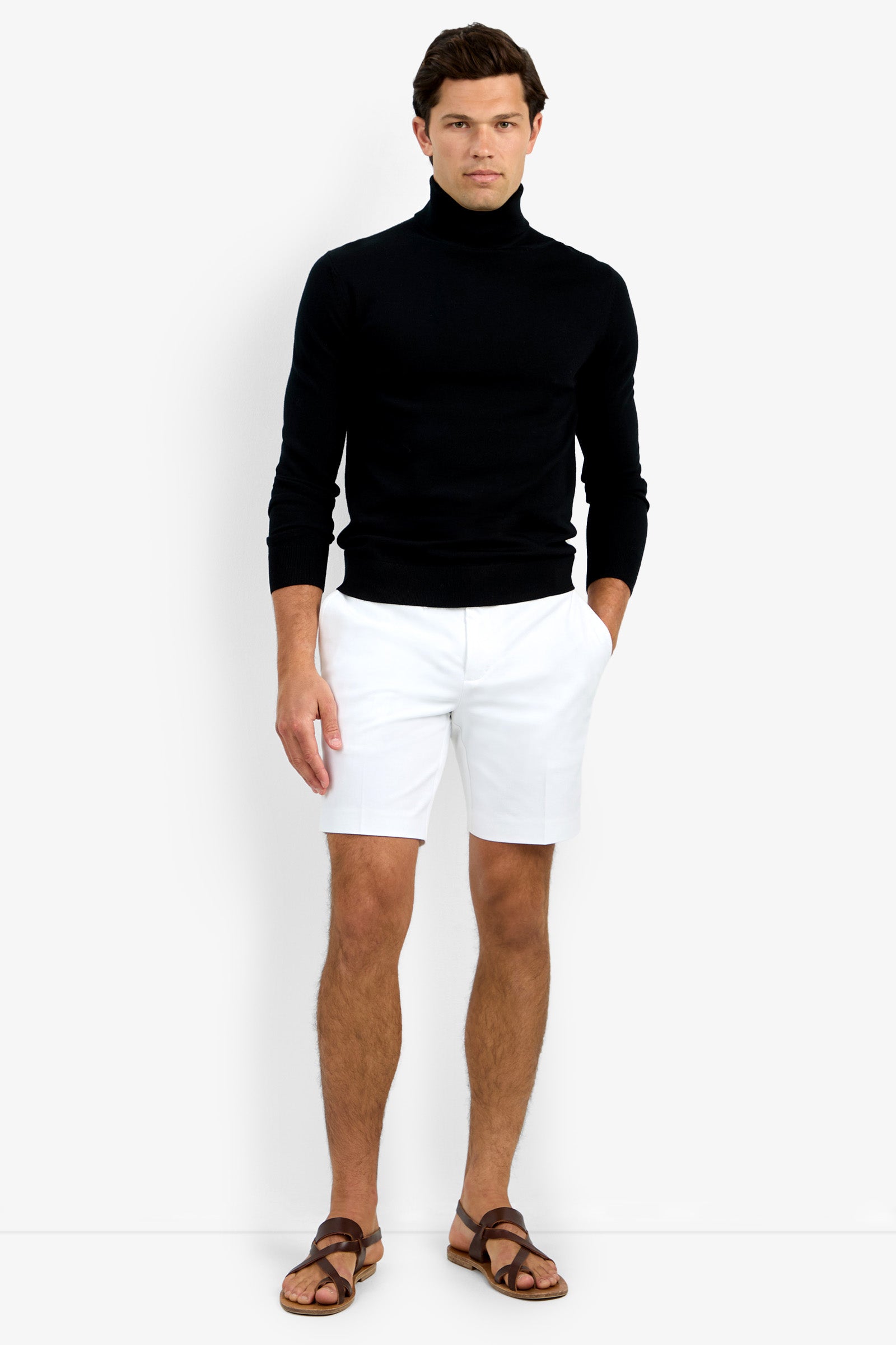 A man stands against a plain background wearing the Roy Long Sleeve Turtleneck in black, white shorts, and brown sandals, with one hand in his pocket.