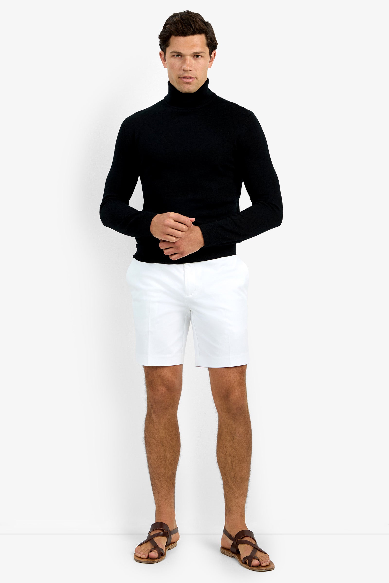 A man with short dark hair stands against a plain white background, wearing the Roy Long Sleeve Turtleneck in black, paired with white shorts and brown sandals. He has a neutral expression and his hands are clasped in front of him.