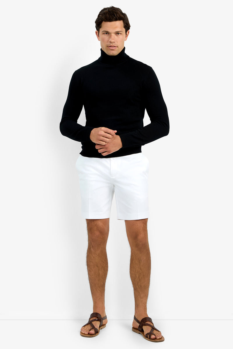 A man with short dark hair stands against a plain white background, wearing the Roy Long Sleeve Turtleneck in black, paired with white shorts and brown sandals. He has a neutral expression and his hands are clasped in front of him.