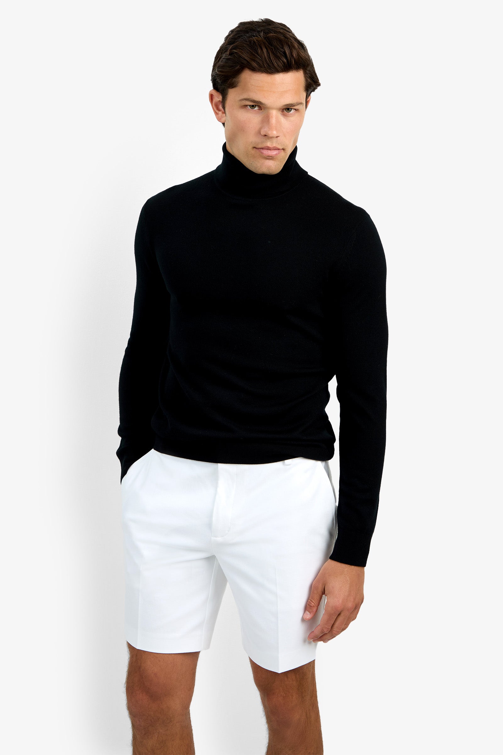 A man confidently poses against a white background, wearing the Roy Long Sleeve Turtleneck in black and white shorts, his left hand by his side and right hand slightly in his pocket.