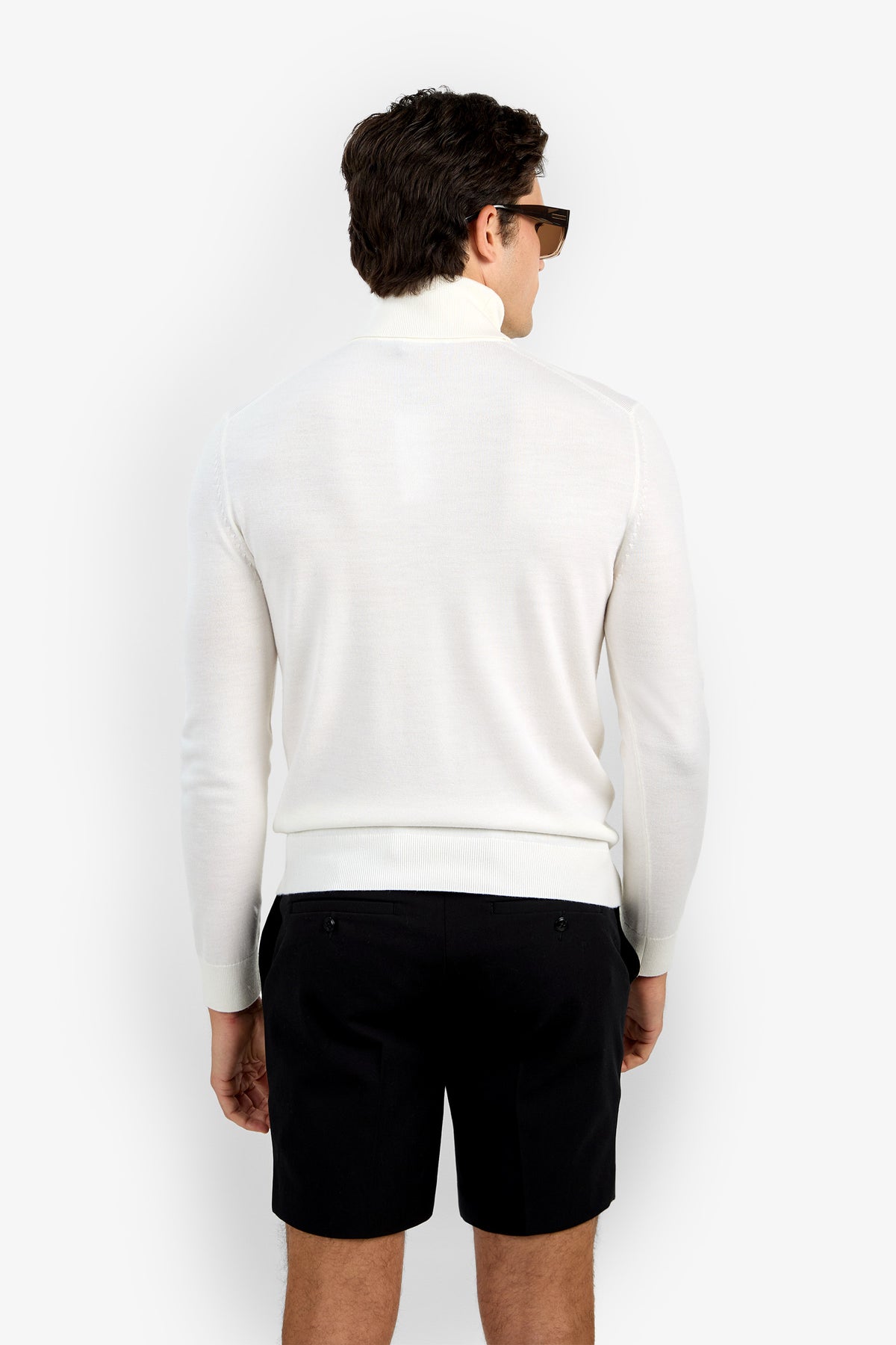 A man with short brown hair, sunglasses, a white Roy Long Sleeve Turtleneck, and black shorts stands facing away from the camera against a plain white background.