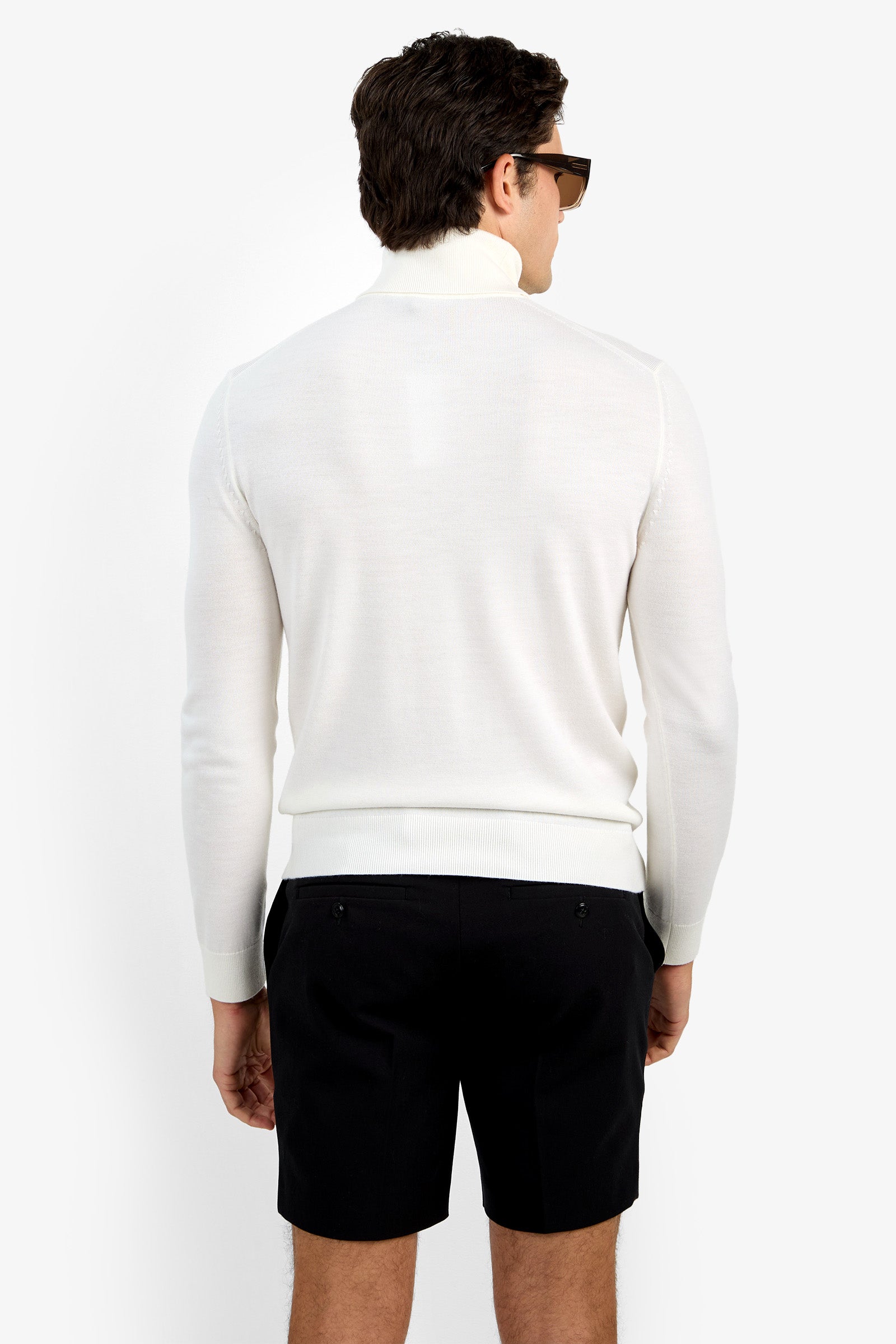 A man with short dark hair and sunglasses, wearing the Roy Long Sleeve Turtleneck in white and black shorts, stands facing away from the camera against a plain white background.