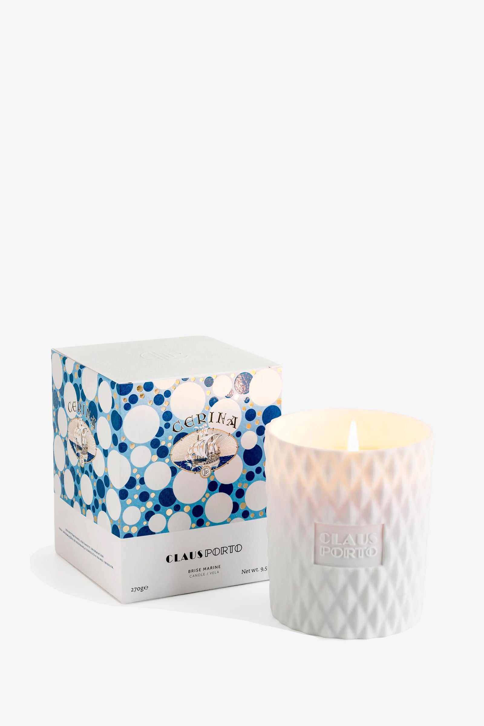 A lit Claus Porto Cerina Candle in a textured glass holder sits next to its blue-and-white patterned box.