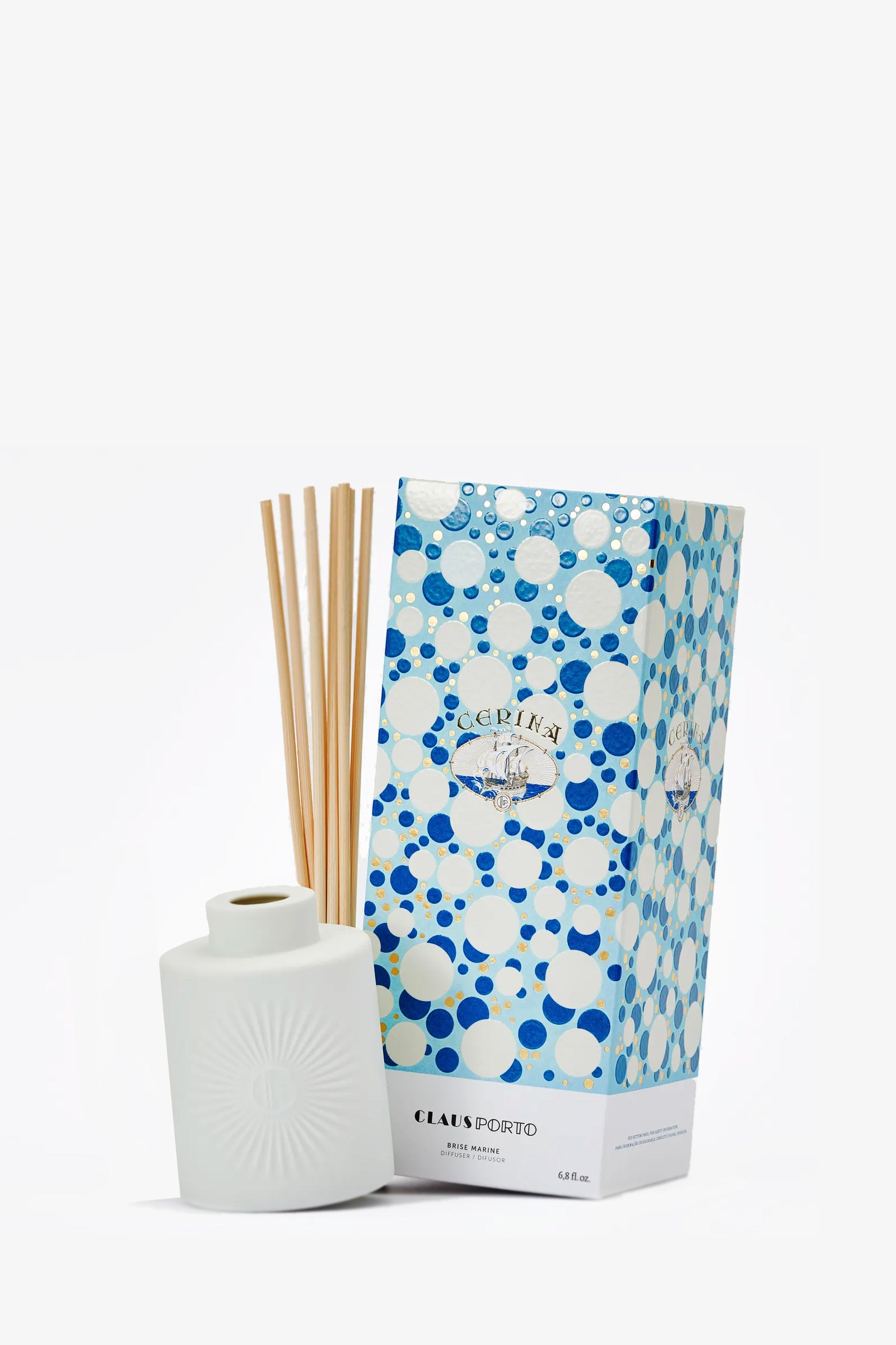 A white ceramic Claus Porto Cerina Diffuser with wooden reeds stands beside a blue and white polka-dotted box featuring circular patterns and elegant branding.