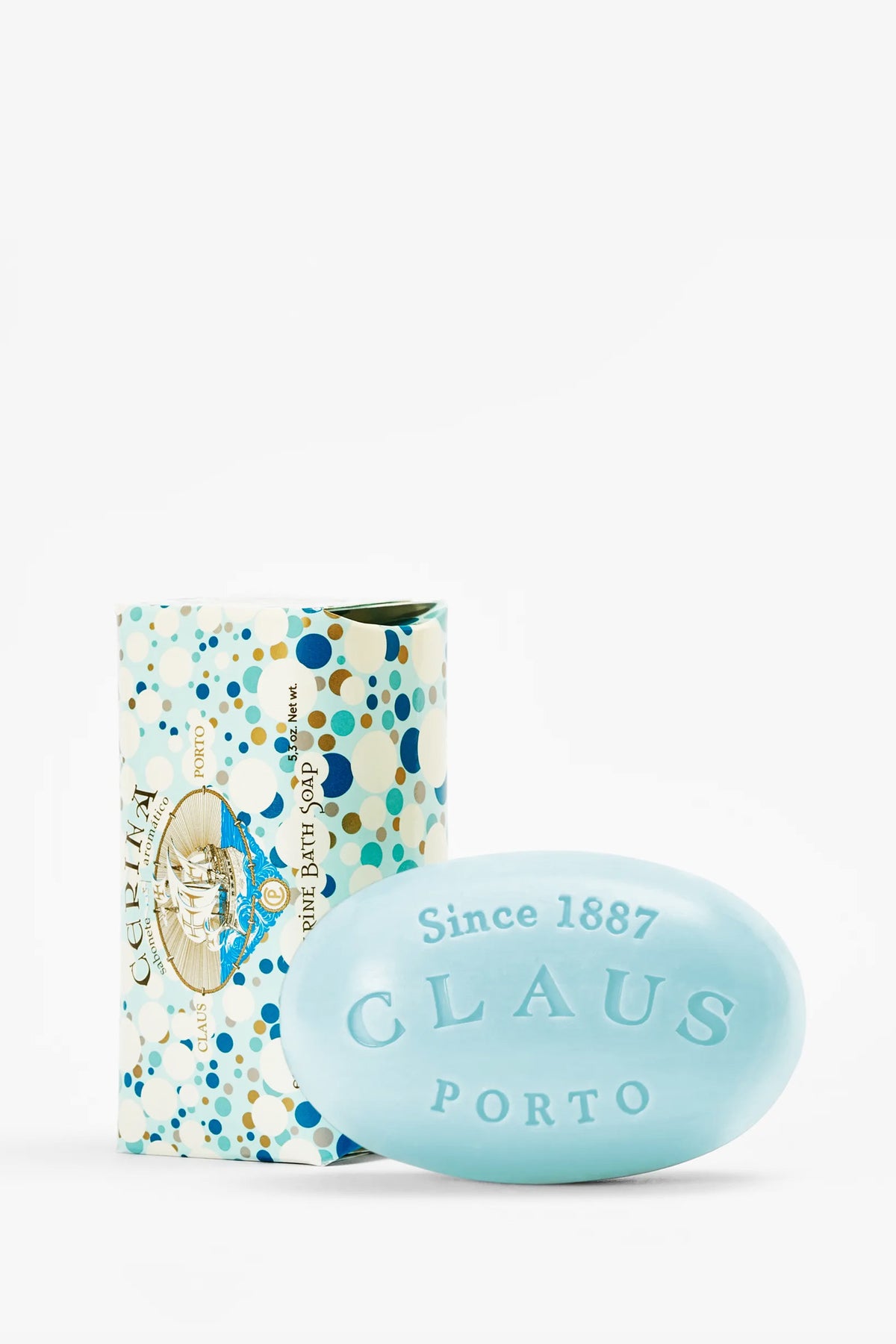 A light blue, oval-shaped Claus Porto Cerina Soap bar labeled “CLAUS PORTO Since 1887” is placed in front of a colorful, patterned soap box featuring blue, gold, and white dots.