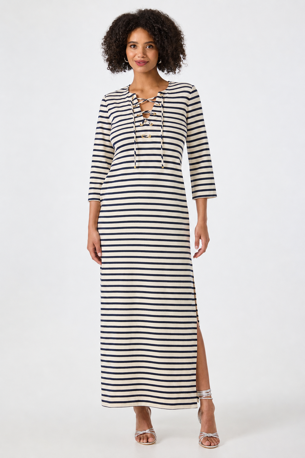 A woman with curly hair stands against a plain white background wearing the Catamaran Dress, a long-sleeve, ankle-length black and white striped dress featuring a lace-up neckline and side slit. She completes her look with silver strappy sandals.