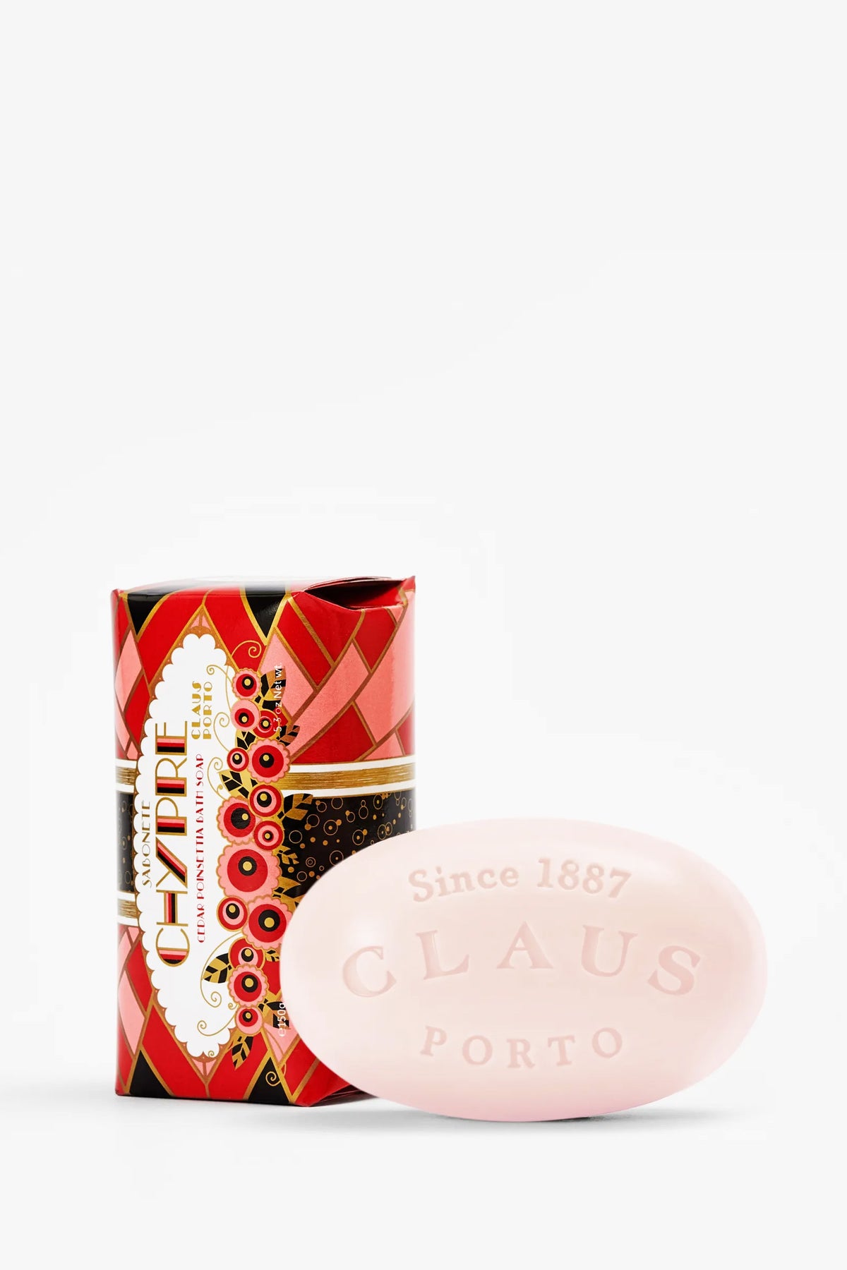 A pink oval Claus Porto Chypre Soap embossed with “Since 1887” sits in front of its decorative red, black, and gold patterned packaging.