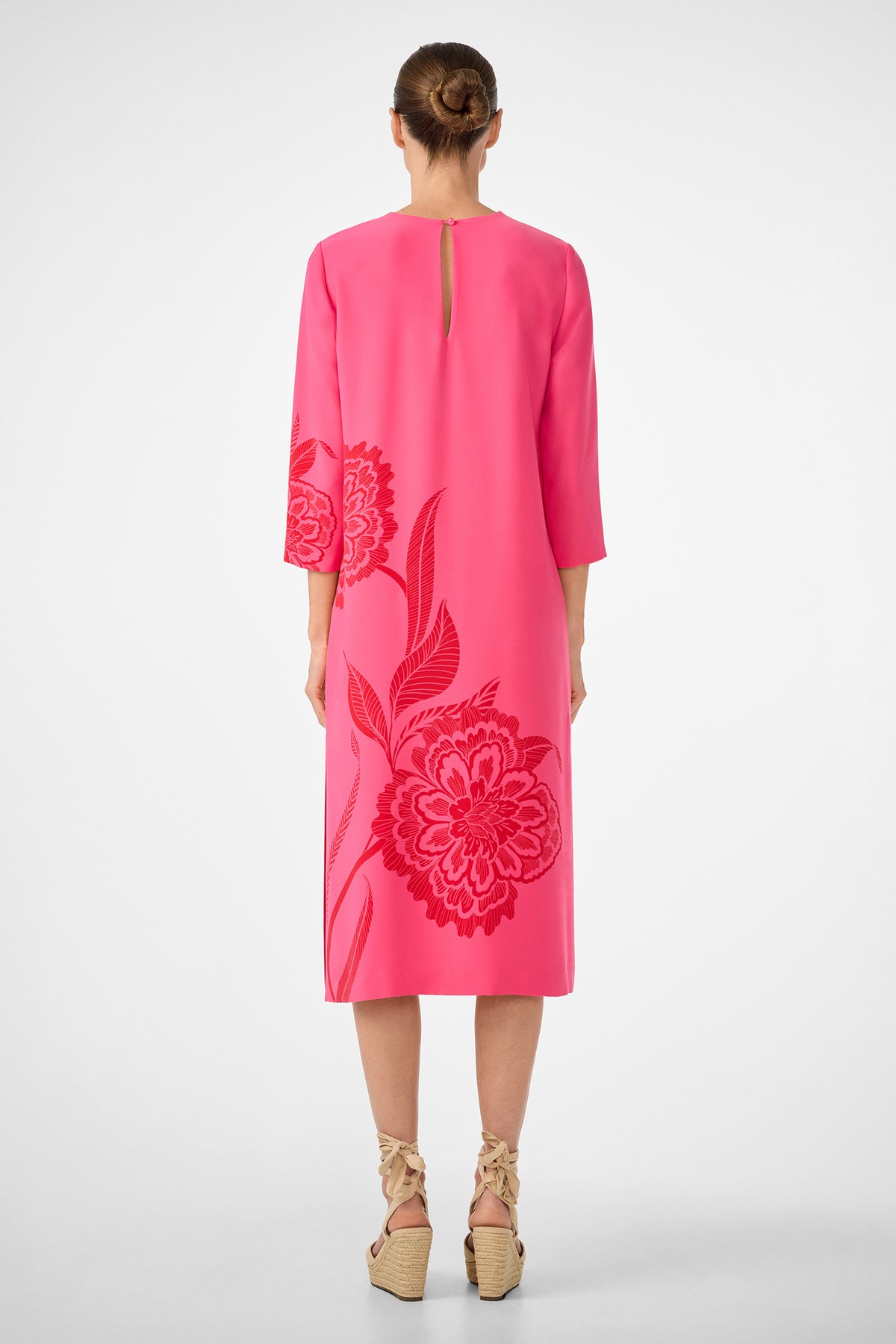 A woman faces away, wearing the Cointreau 2 Dress—a bright pink midi dress with floral patterns, elbow-length sleeves, and a small back keyhole detail. Her hair is in a bun, and she pairs the look with beige wedge sandals featuring ankle ties.