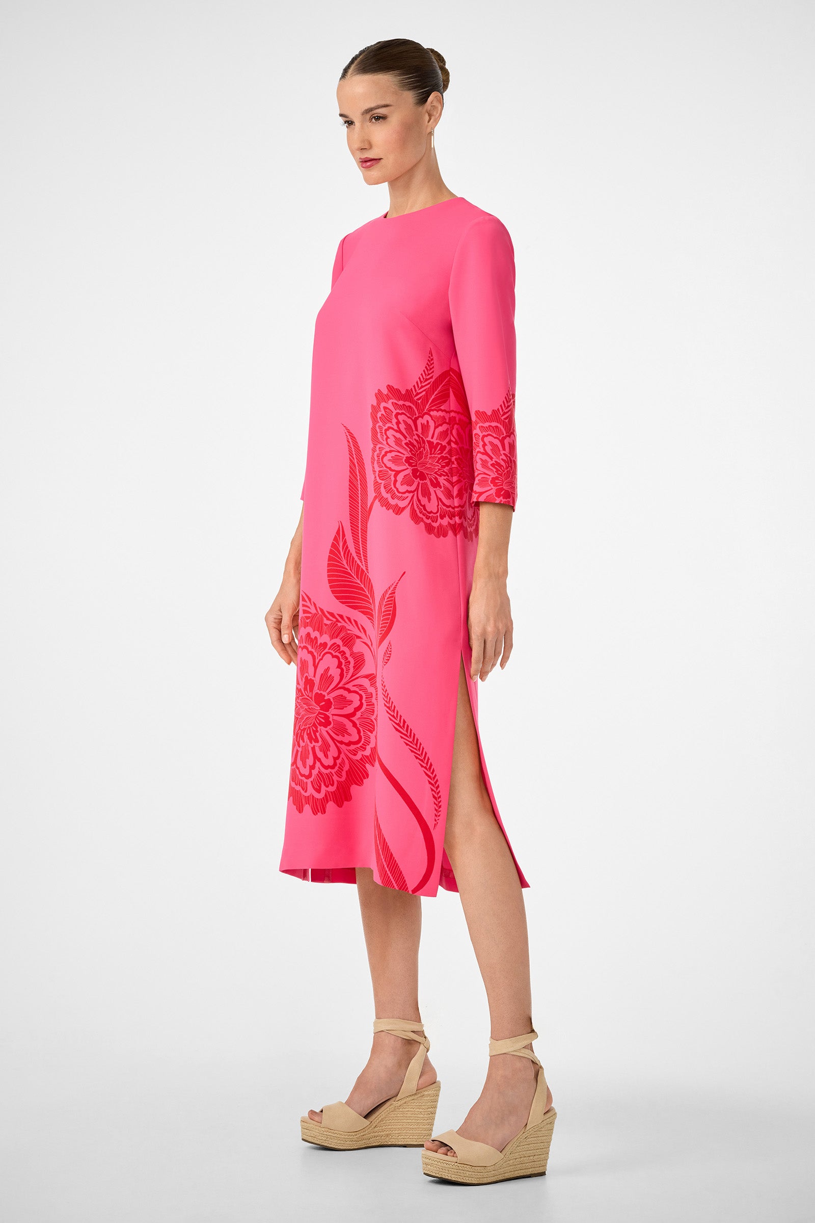 A woman in profile wears the Cointreau 2 Dress, a bright pink knee-length dress with floral patterns and three-quarter sleeves. Her hair is styled in a sleek bun and she pairs the look with beige wedge sandals.