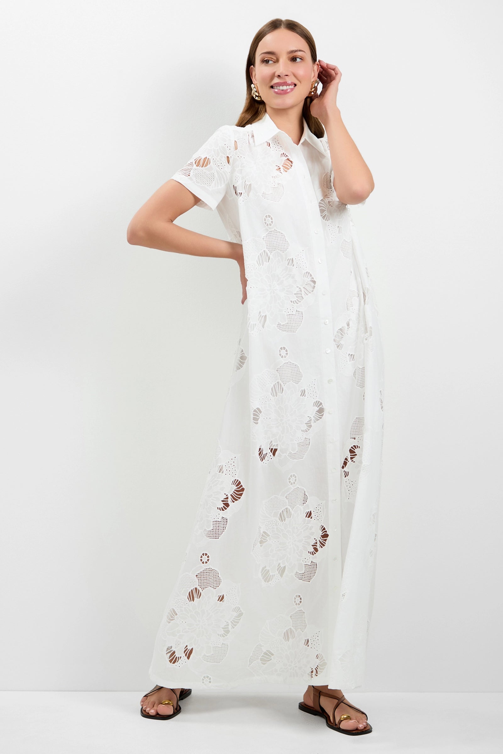 A woman smiles and stands against a white background, wearing the Kon Tiki Maxi Dress—a long, white, semi-sheer dress with floral cutout patterns and short sleeves—paired with sandals. She touches her ear with one hand.