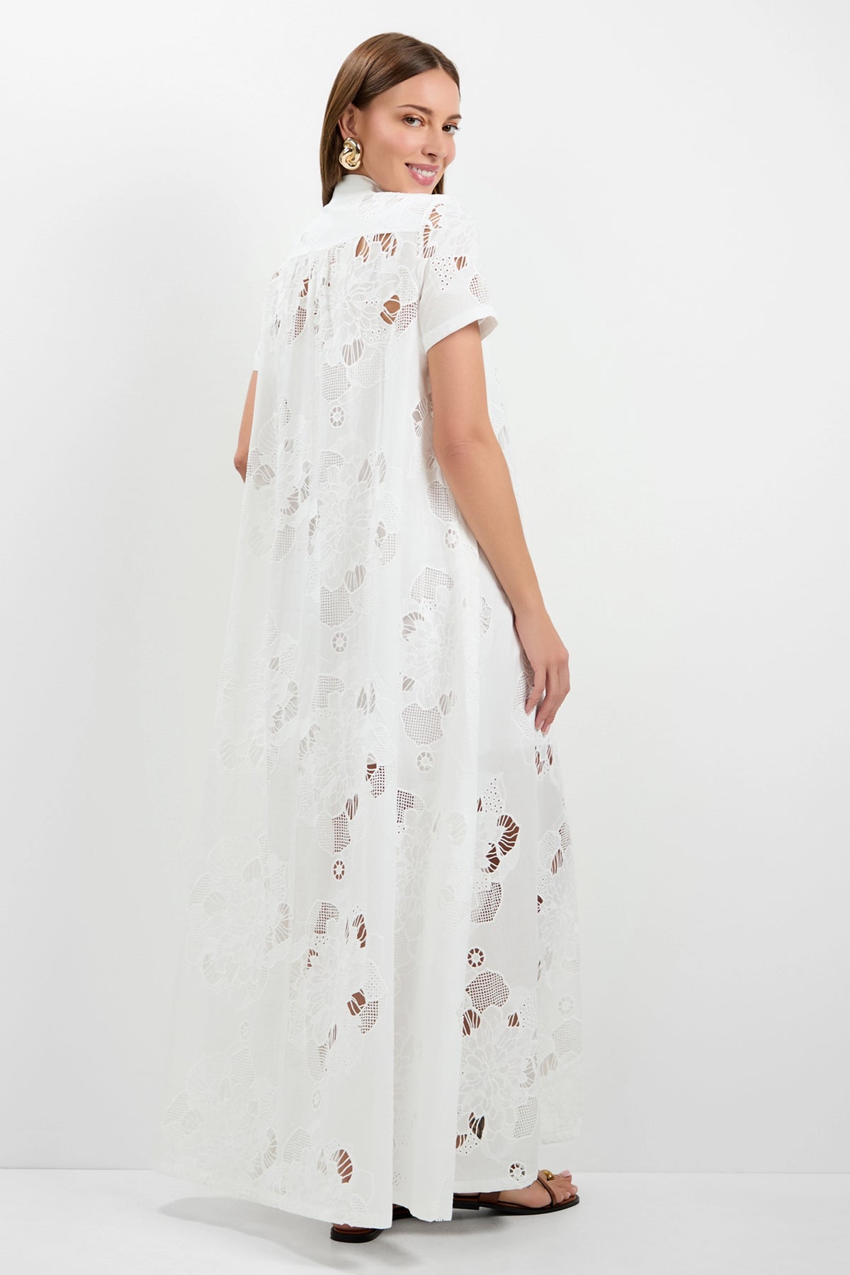 A woman with straight brown hair, gold earrings, and black sandals stands against a white background, looking over her shoulder and smiling while wearing the Kon Tiki Maxi Dress—a long, white, sheer floral lace dress with short sleeves.