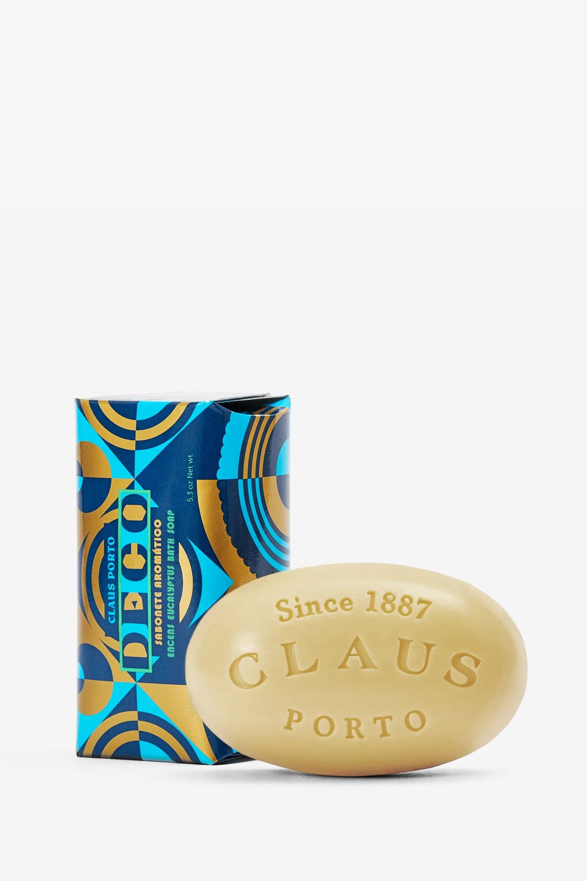 A beige, oval-shaped Claus Porto Deco Soap bar featuring “Since 1887 CLAUS PORTO” embossed on its surface, positioned in front of its blue and gold geometric-patterned box labeled “Deco.”.