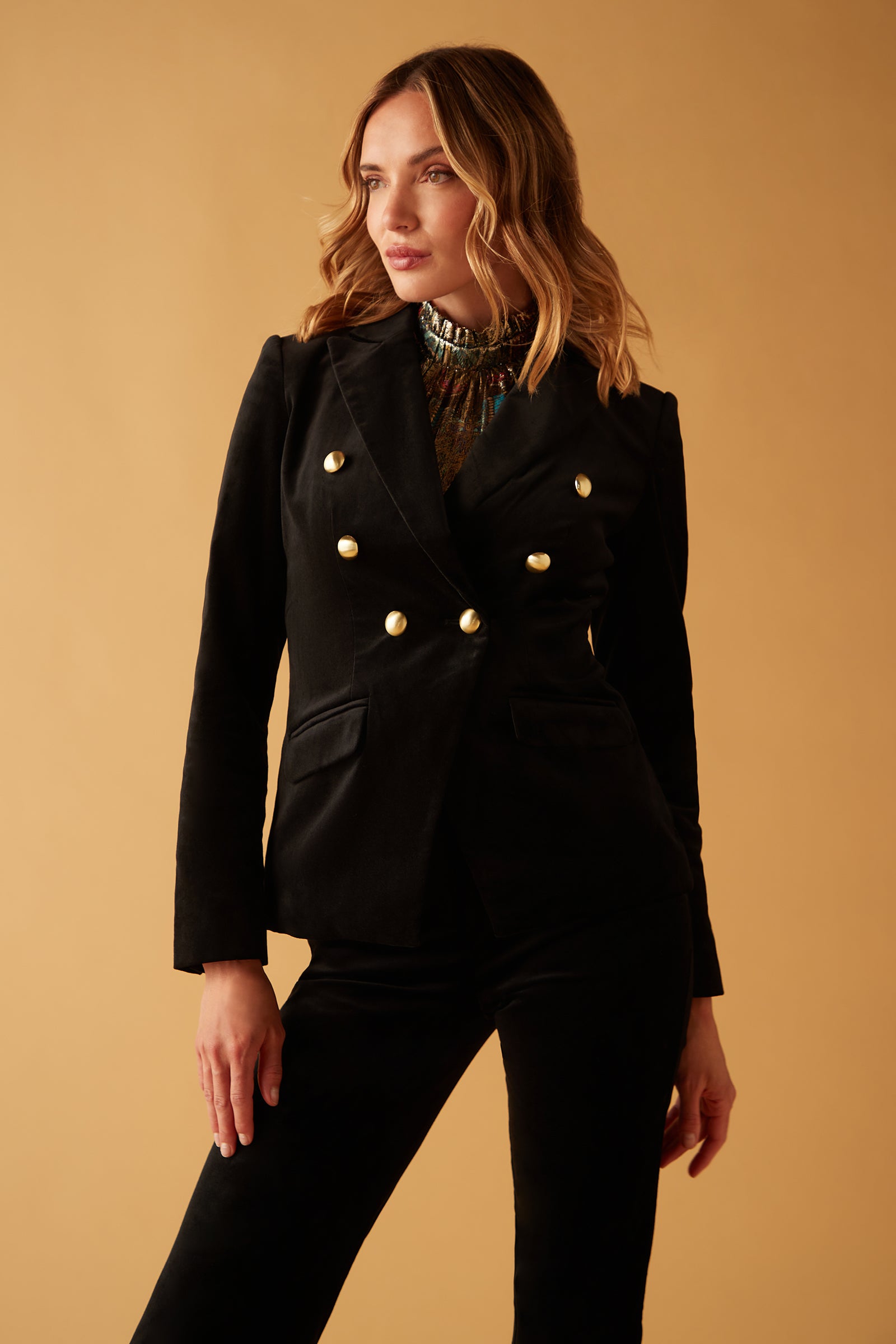 A woman with wavy blonde hair stands against a beige background, wearing the fitted black Midlands Velveteen Blazer Jacket with gold buttons and matching black pants, gazing slightly to the side.
