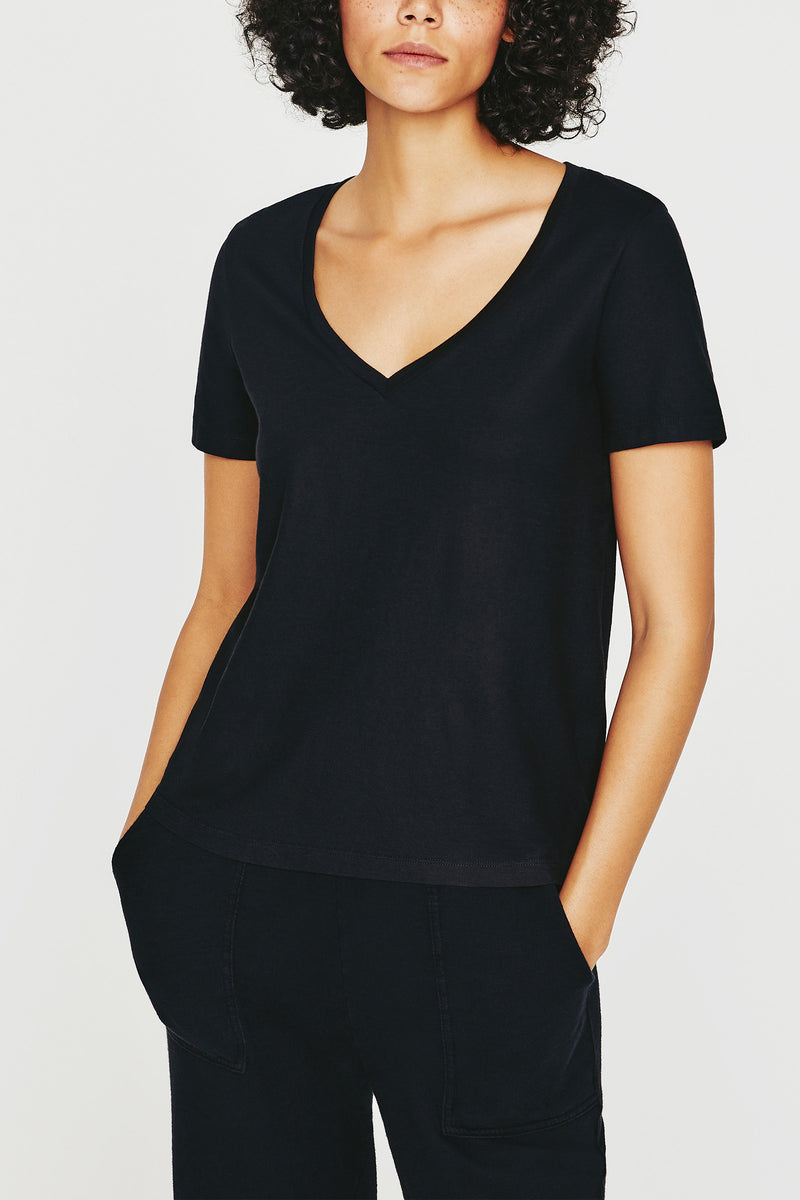 A person with curly hair wears the AG Jager Vee Tee in black and black pants, standing hands-in-pockets against a white background. Their face is partially out of frame.