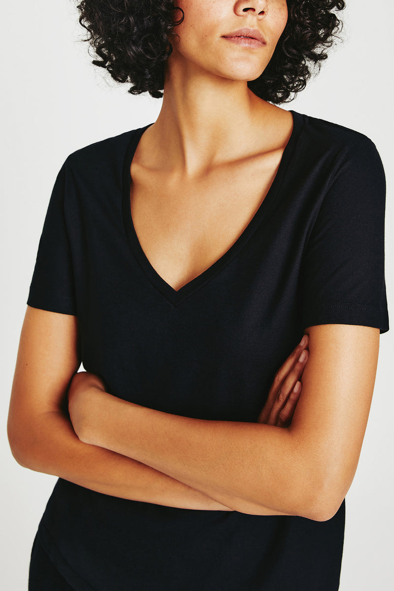 A woman with curly hair wears the AG Jager Vee Tee in black, standing with arms crossed against a white background.