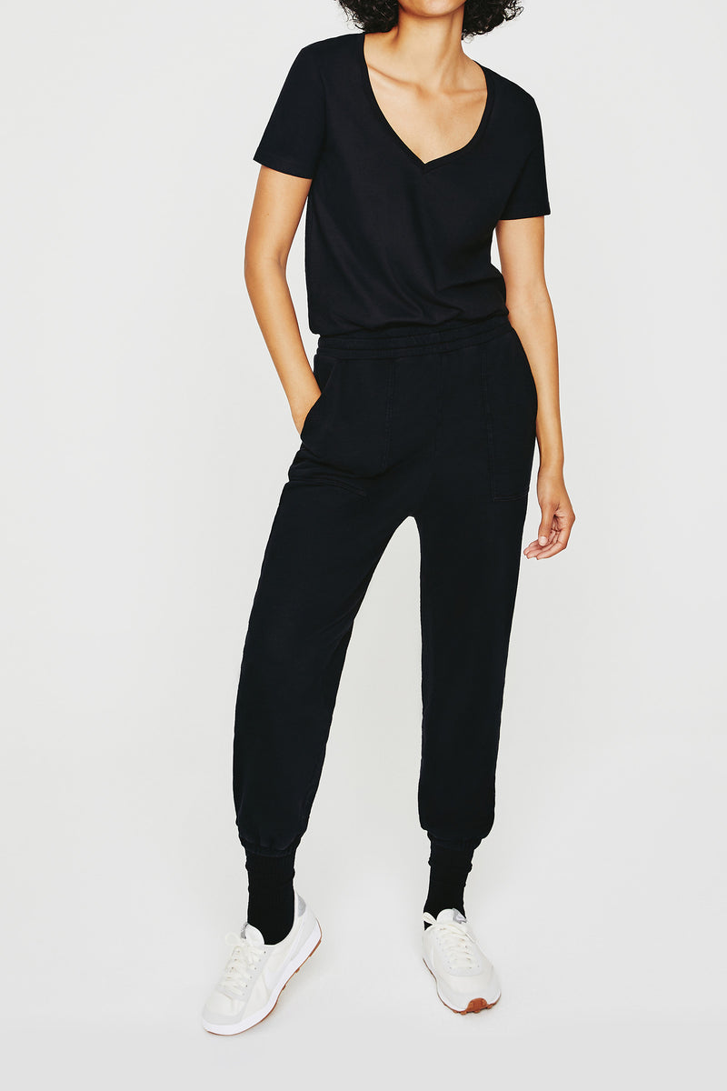 A person wearing the AG Jager Vee Tee in black, black jogger pants, and white sneakers stands with hands in pockets against a plain white background.