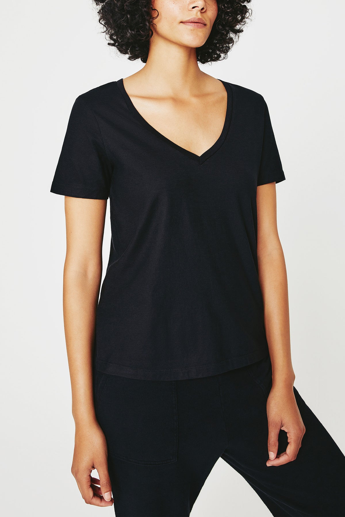 Someone wearing the AG Jager Vee Tee - Black and black pants stands against a plain white background, shown from shoulders to hips.