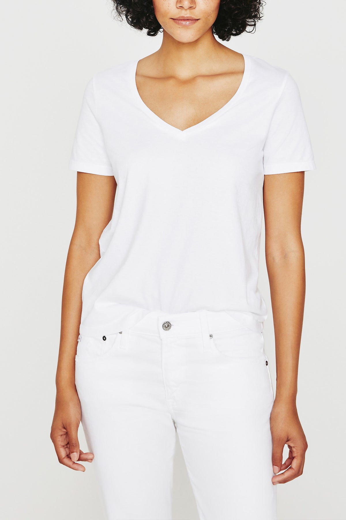 A woman poses against a plain background wearing the AG Jager Vee Tee in white, paired with white jeans. Her head is slightly tilted, and only her upper body is shown.