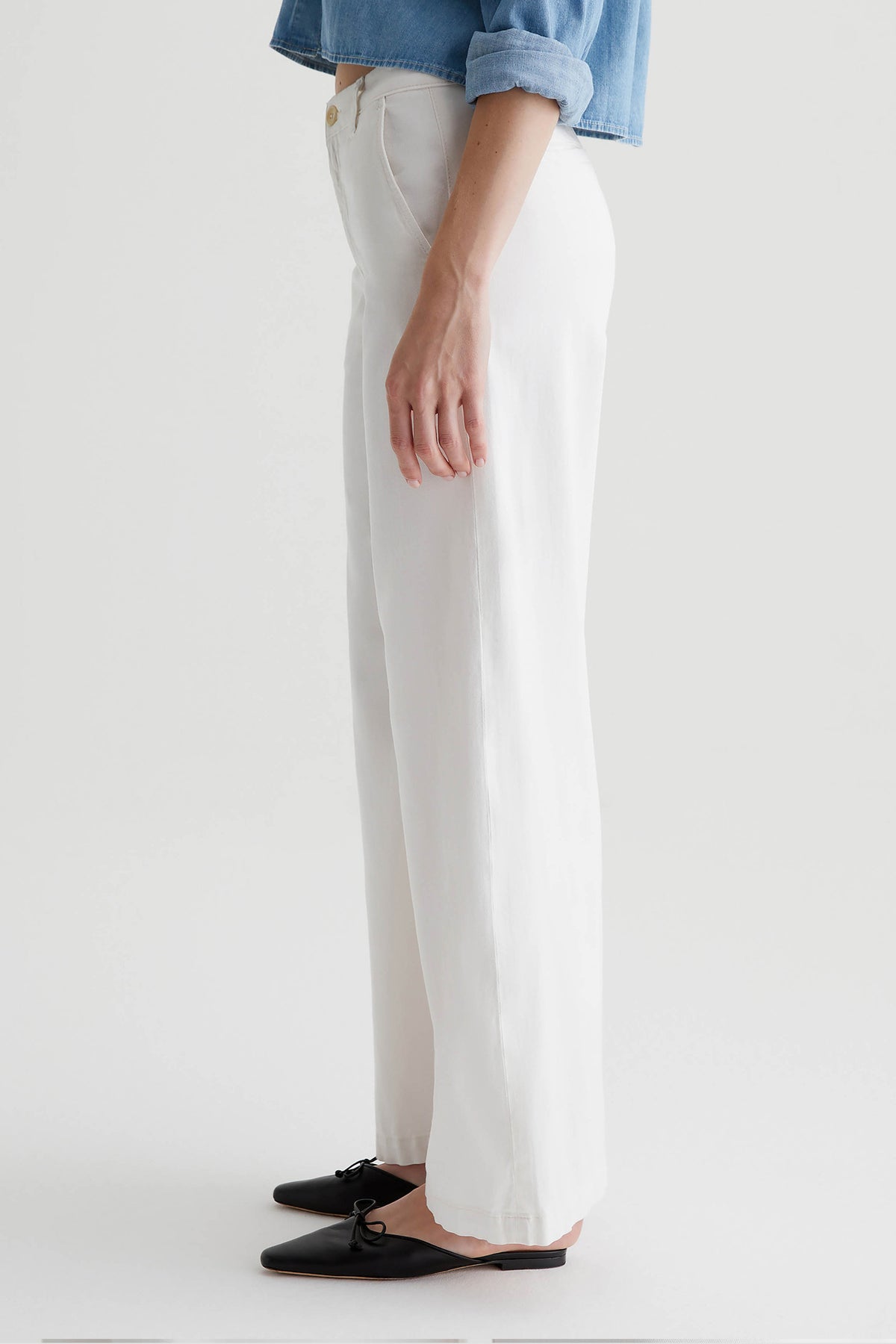 A person stands in profile wearing the AG Caden Wide Leg Pant in Powder, a light blue rolled-sleeve shirt, and black flat shoes with a small bow. The background is plain and light.
