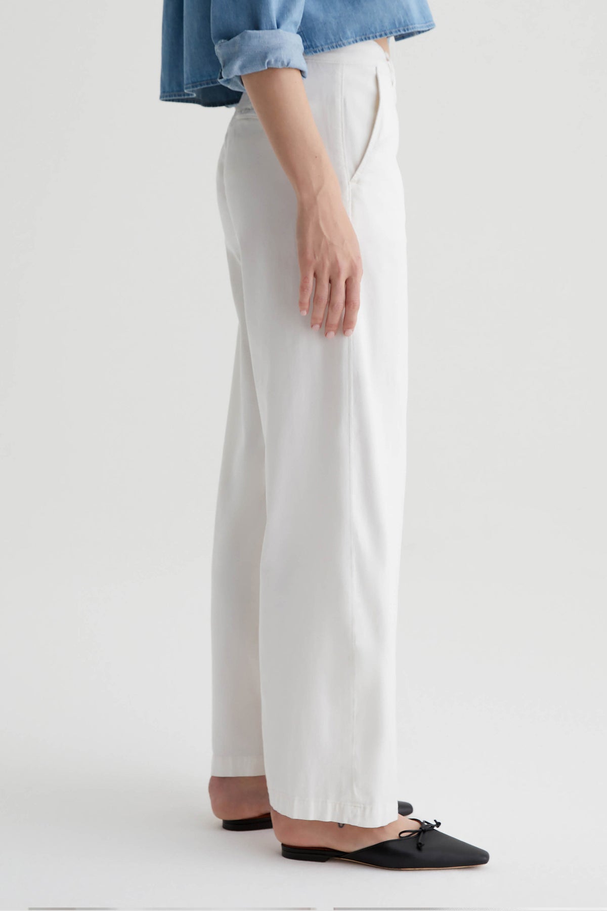 A person stands in profile against a light background, wearing the AG Caden Wide Leg Pant in Powder, a light blue cuffed shirt, and black pointed-toe flats with bows.