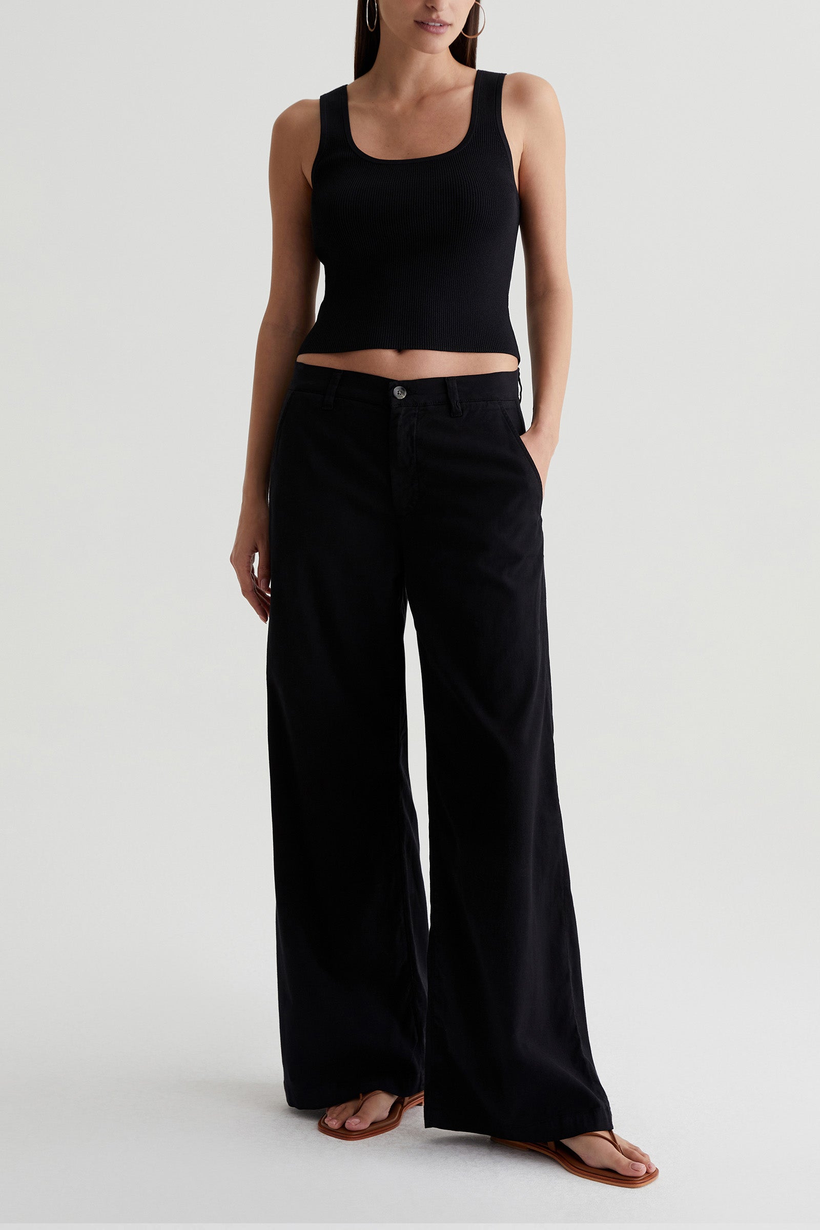 A person stands against a light background wearing the AG Caden Wide Leg Pant in black, paired with a black sleeveless crop top and brown flip-flops. Their hands are in their pockets, and their face is not visible.