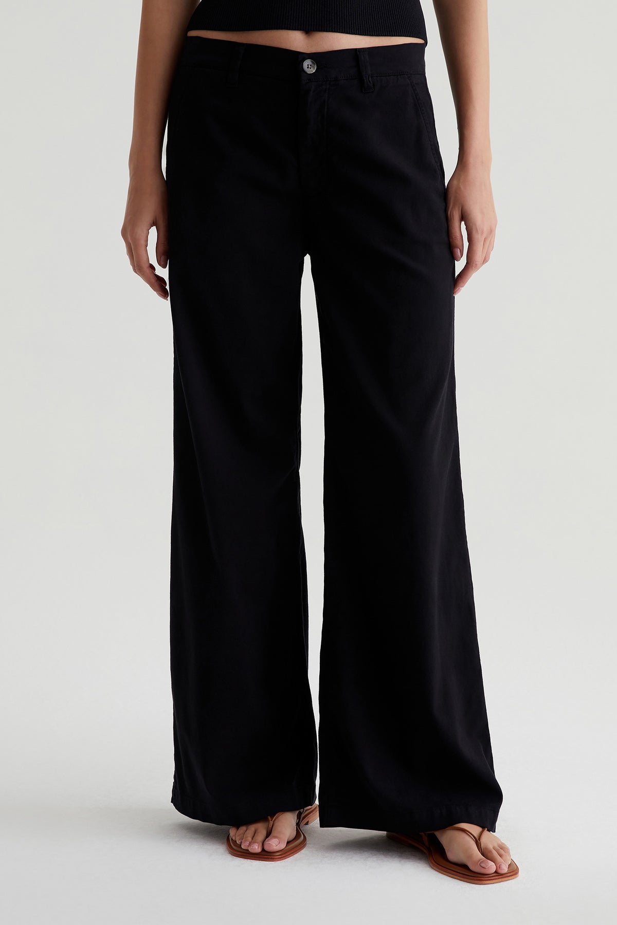 A person wearing AG Caden Wide Leg Pant in black and brown sandals stands against a plain light background, showing only the lower torso and legs.