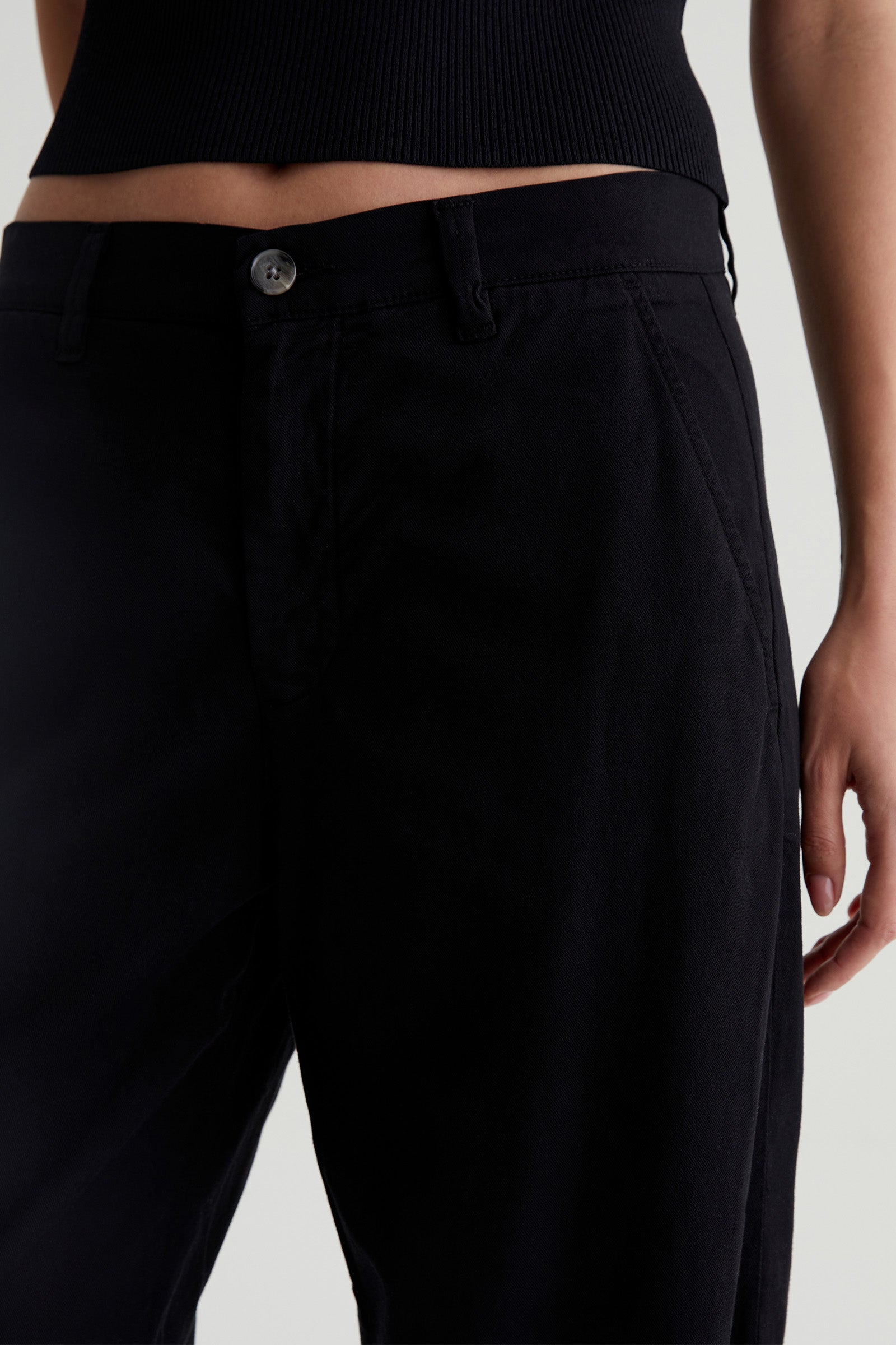 Wearing the AG Caden Wide Leg Pant in black, shown from waist to mid-thigh, paired with a black ribbed top and one hand resting by their side.