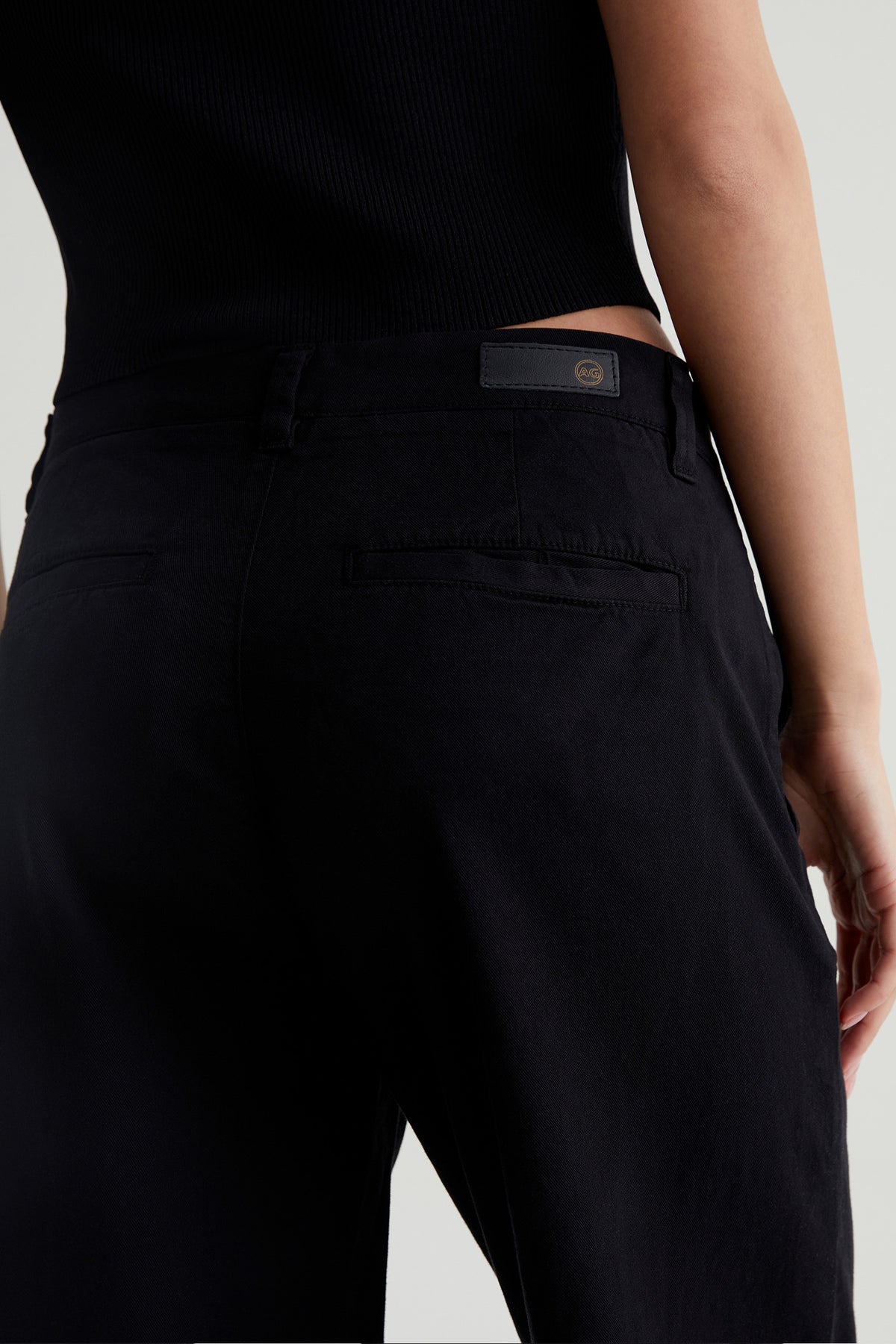 A person wearing the AG Caden Wide Leg Pant in black, shown from the waist down and behind, with one arm relaxed at their side.