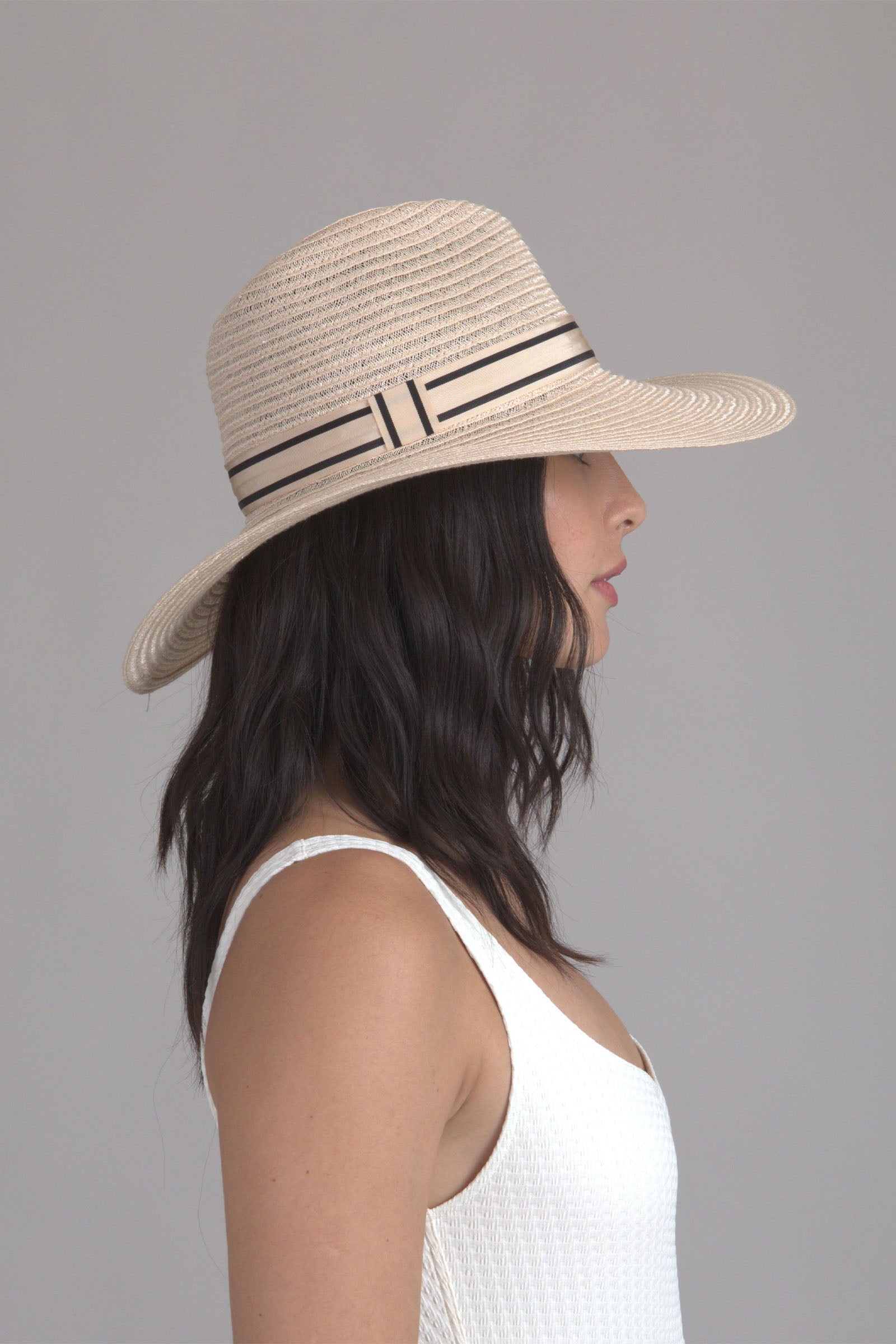 A woman with wavy dark hair wears the Eugenia Kim Emmanuel Sun Hat, featuring a wide brim and black and beige band, paired with a sleeveless white textured top, shown in profile against a plain light gray background.