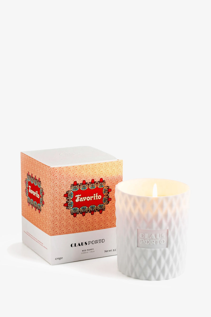 A lit Claus Porto Favorito Candle in a textured glass holder sits next to its decorative box.