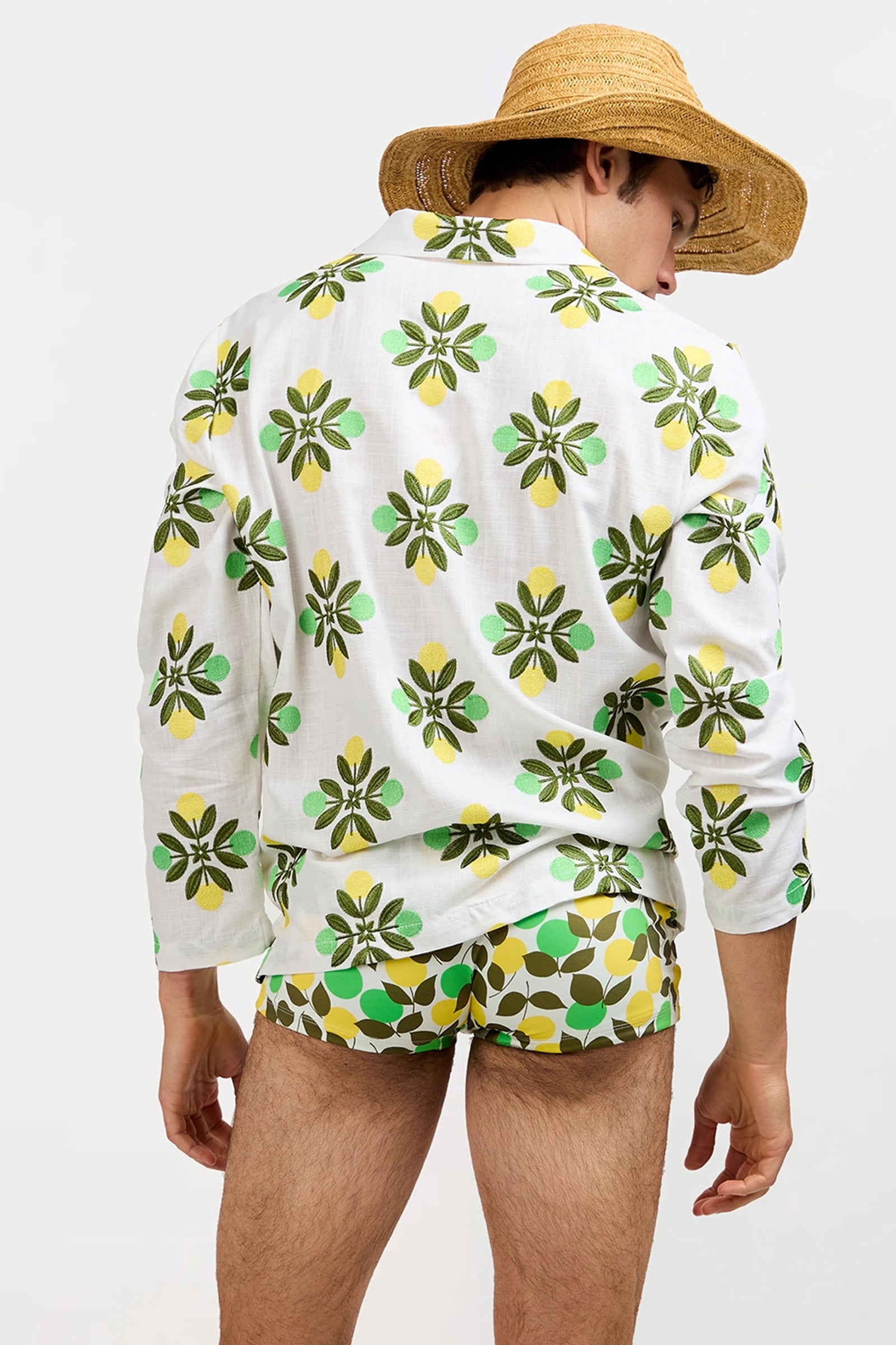 A man stands with his back to the camera against a plain light background, wearing the Holmes Pullover, a white shirt featuring a green and yellow floral pattern, paired with green patterned swim trunks and a straw hat.