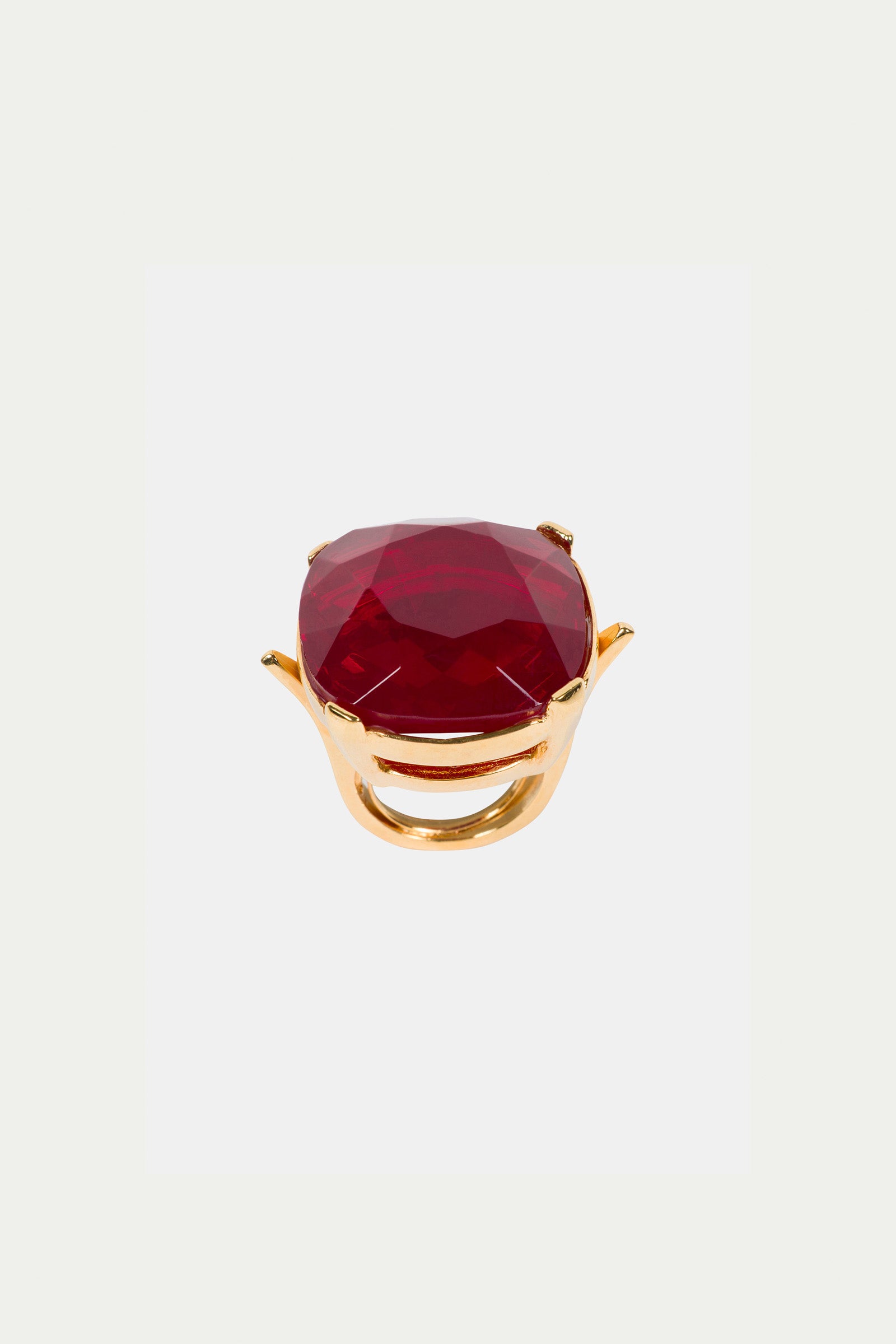 The Ruby Faceted Cocktail Ring showcases a large, faceted oval red gemstone in a raised prong setting, elegantly presented against a plain white background.