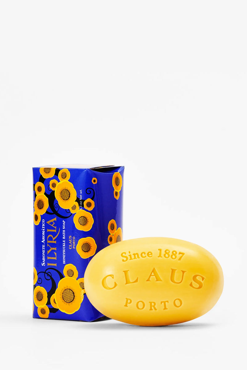 The Claus Porto Ilyria Soap, a yellow oval bar engraved with “Since 1887 CLAUS PORTO,” is elegantly displayed in front of its blue box adorned with yellow and orange floral patterns. This natural honeysuckle soap rests gracefully on a plain white background.