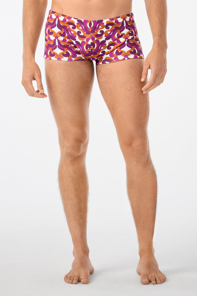 A person stands barefoot on a plain background, wearing Jet Set Swim Trunk - Dewdrop with vibrant purple, orange, and white patterns. The persons head is not shown in the image.