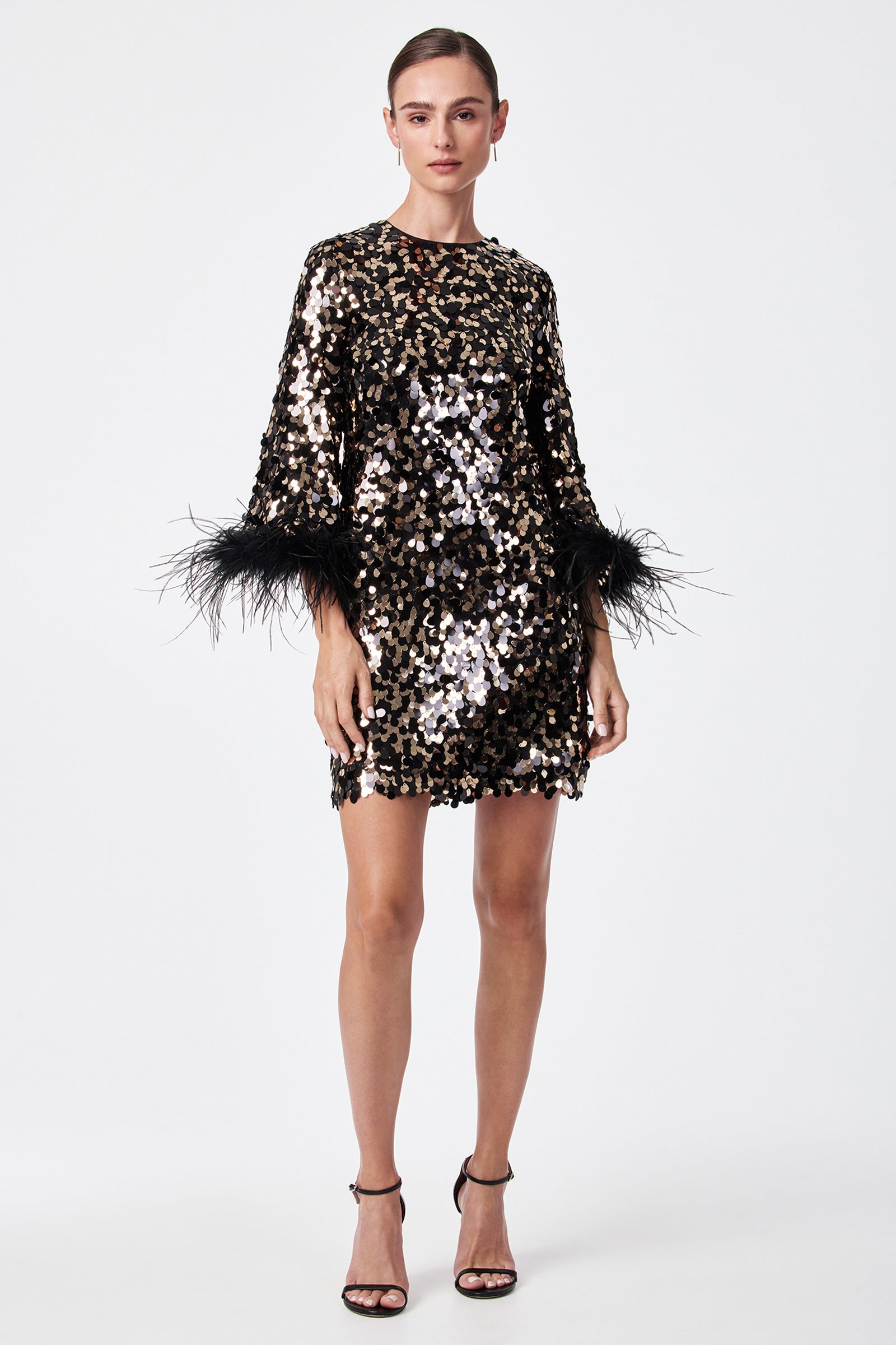 A woman stands against a plain white background wearing the Juliet Dress—a short, long-sleeved black and gold sequin dress with feathered cuffs—paired with black high-heeled sandals.