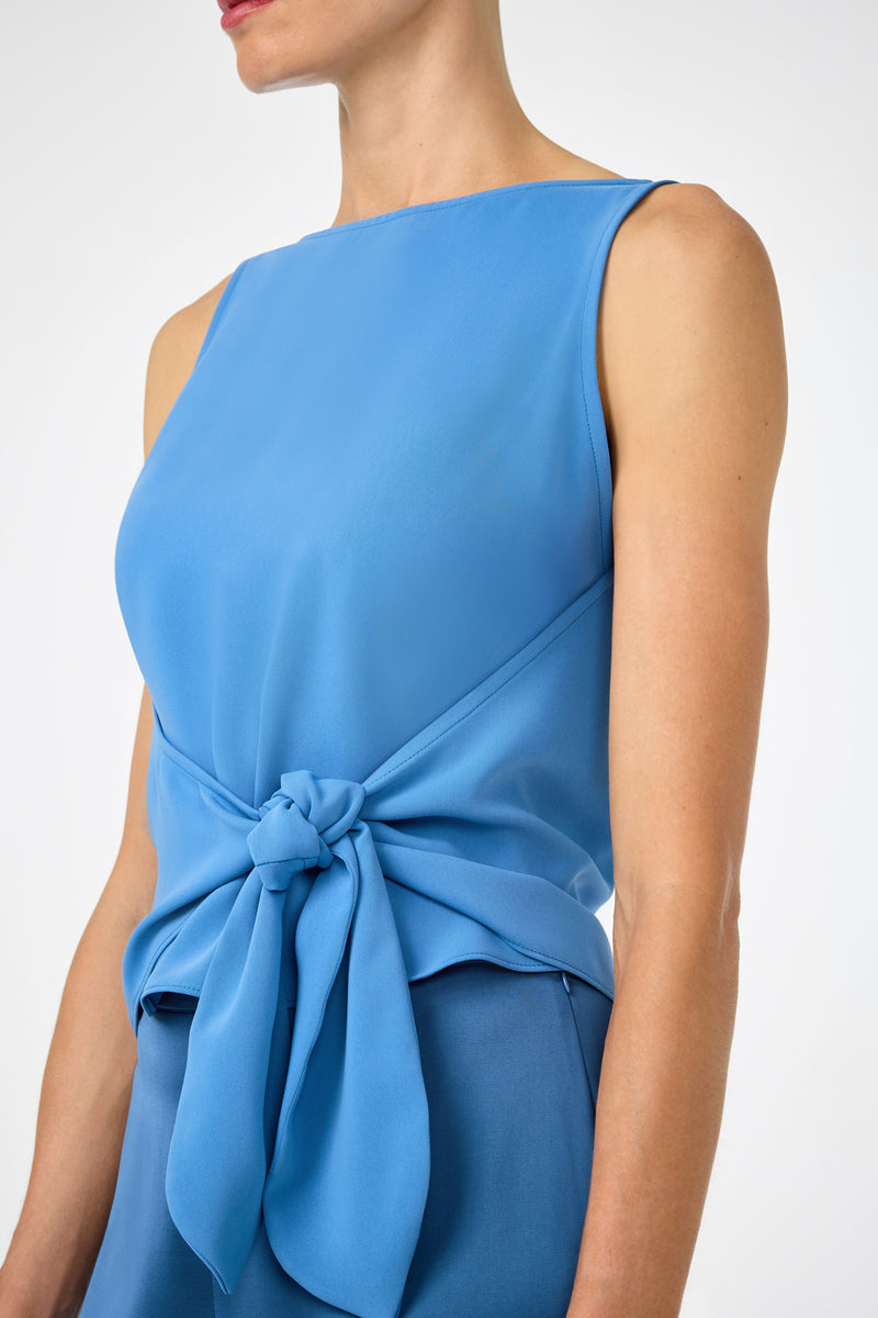 A woman wears the Kuri Tie-Front Top, a sleeveless, boat-neck blue top with a waist tie detail, paired with matching blue pants. Her face is partially out of frame.