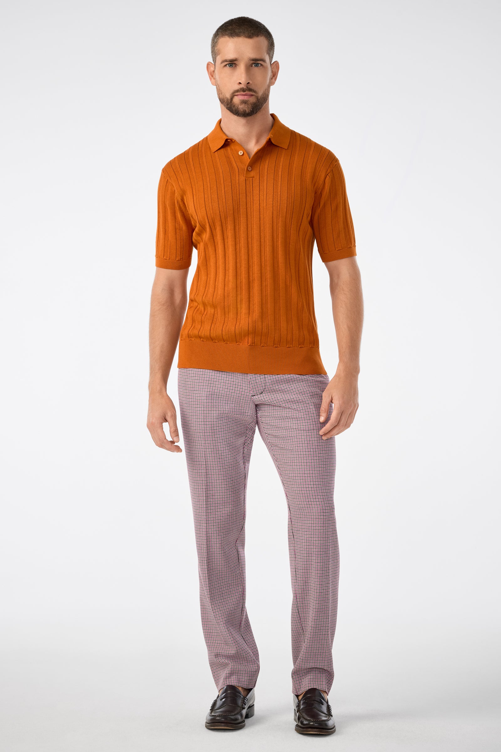 A man with short hair and a beard stands against a plain background wearing the Kensington Short-Sleeve Polo in Geranium + Amber, light gray plaid pants, and dark brown dress shoes. His expression is neutral.