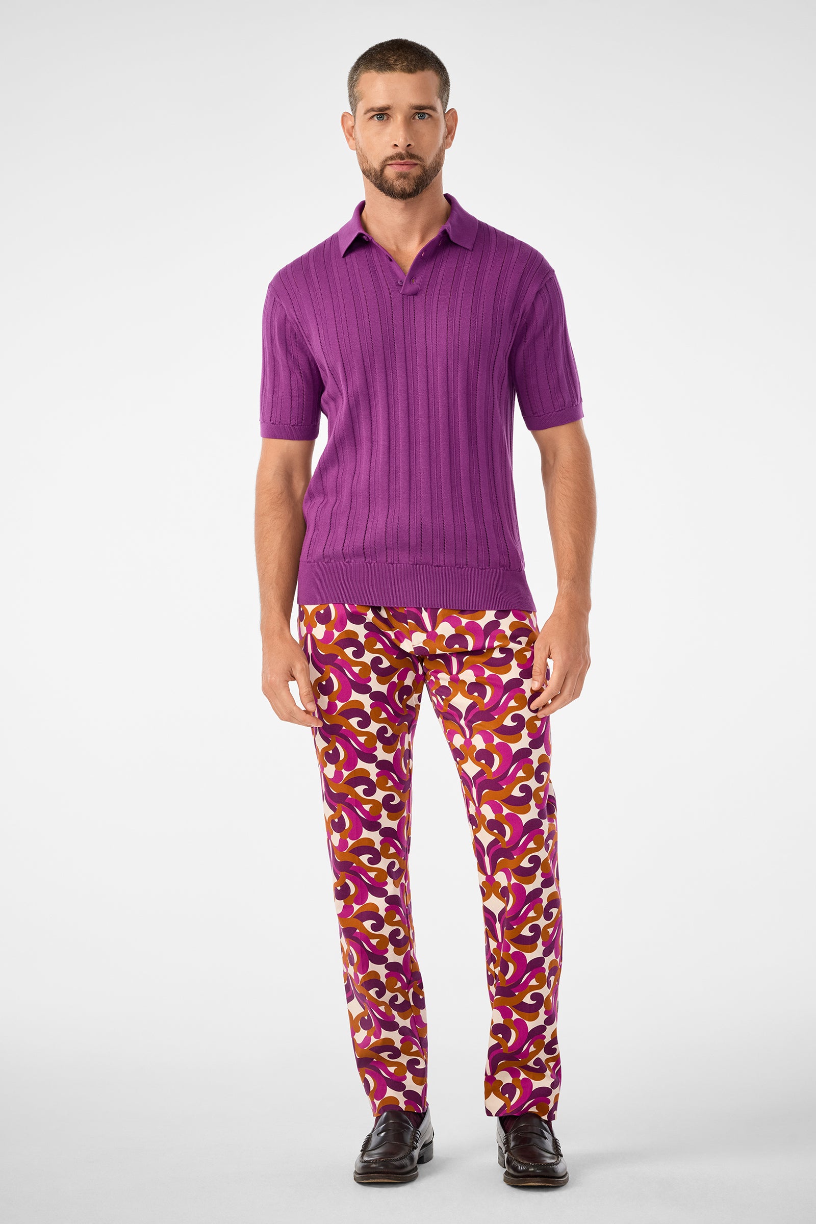 A man with short hair and a beard stands against a plain background in the Kensington Short-Sleeve Polo (Geranium + Amber) and vibrant red, yellow, and white patterned pants, paired with dark shoes.
