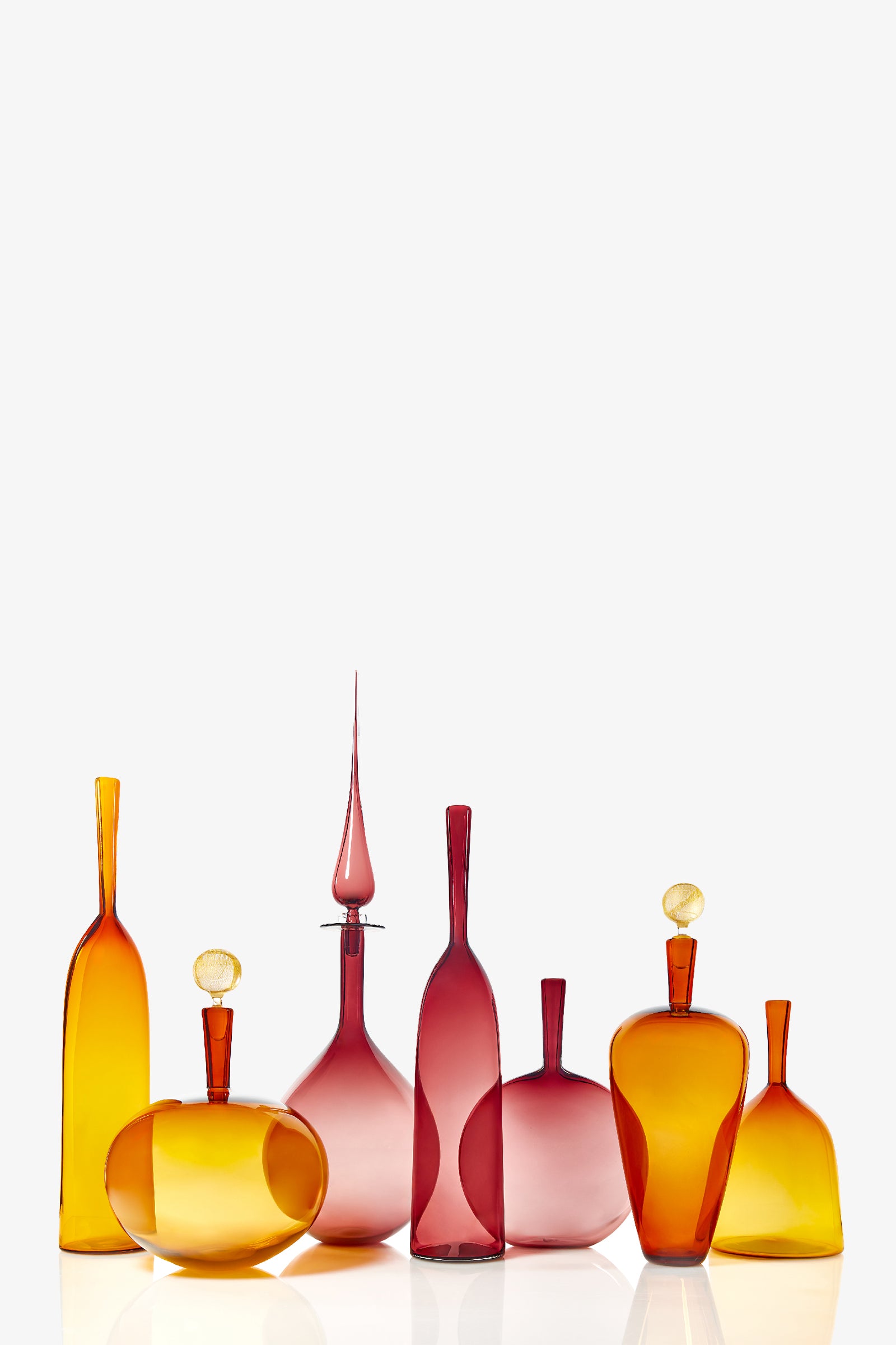 A Large Angelic Bottle, in warm amber, orange, or pink glass with a decorative stopper, stands among seven uniquely shaped and sized bottles arranged side by side against a plain white background.