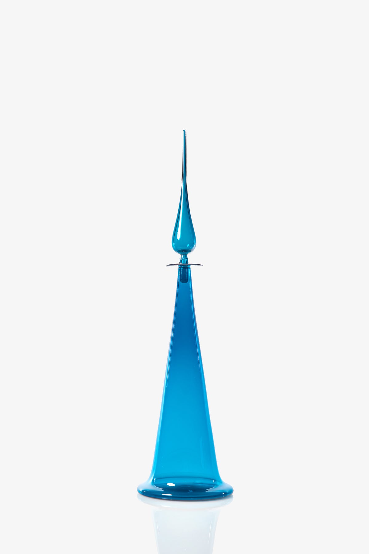 The Large Fluted Cone Decanter, a tall and slender blue glass bottle with a pointed stopper, sits on a reflective white surface against a plain white background.