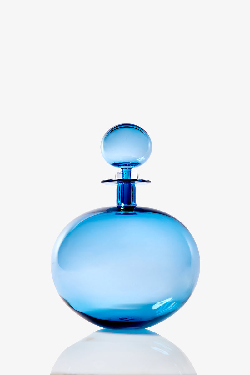 A Large Low Round Decanter made of blue glass with a matching stopper sits on a reflective surface against a plain white background.