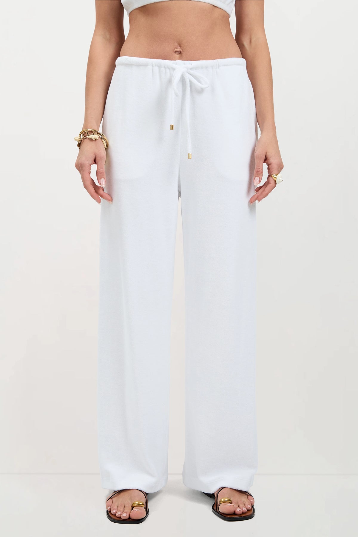 A person wearing Linsk Wide-Leg Pant - Poolside Terry, a cropped white top, gold bracelets, rings, and black sandals stands against a plain light background.