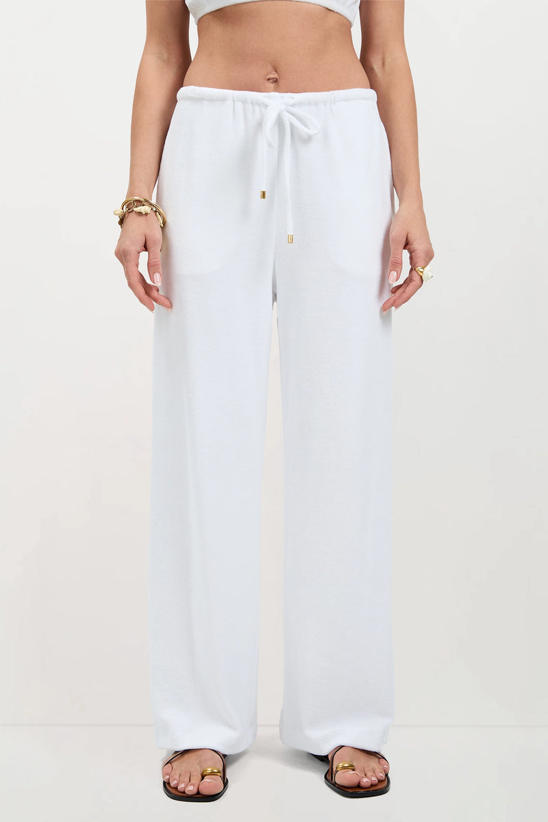 A person wearing Linsk Wide-Leg Pant - Poolside Terry, a cropped white top, gold bracelets, rings, and black sandals stands against a plain light background.