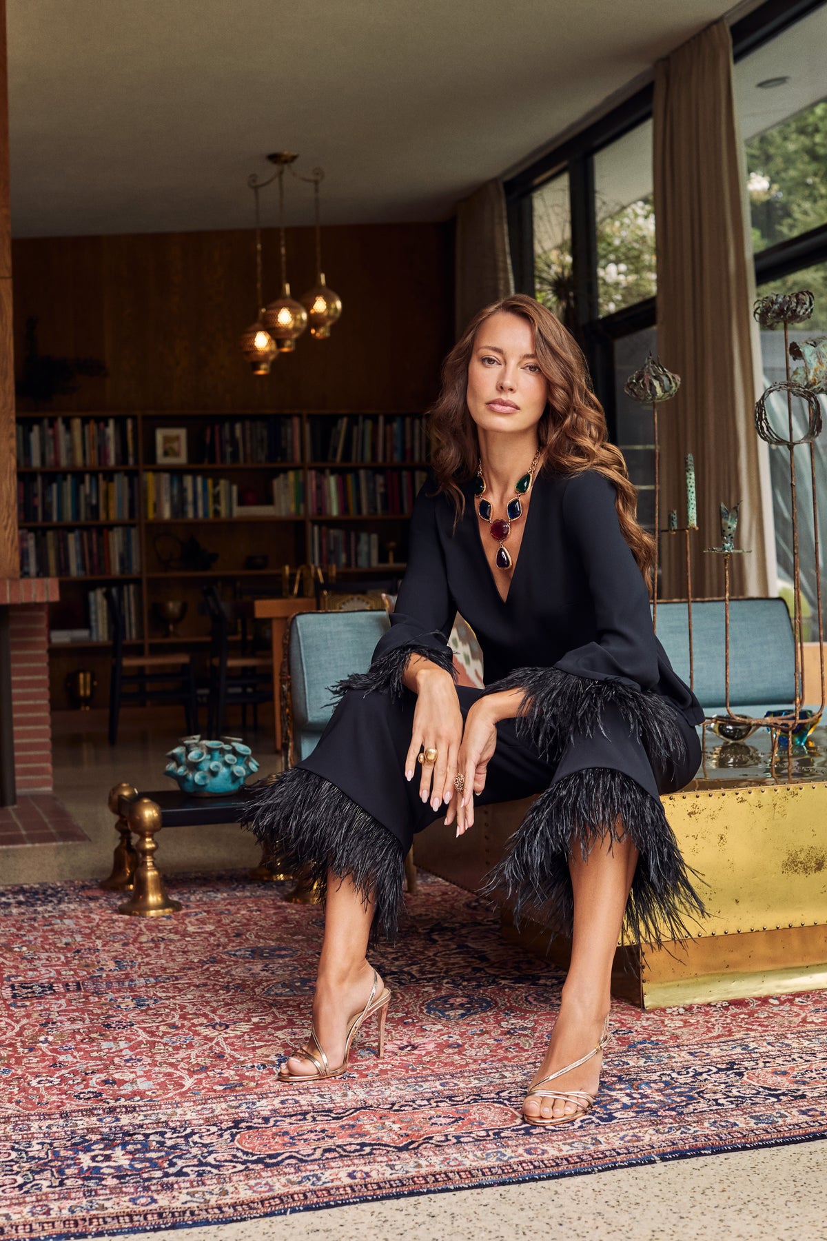 A woman sits wearing the black Anina Top and matching cropped trousers, both trimmed with dark feathers, paired with gold strappy heels and a large multi-colored statement necklace.