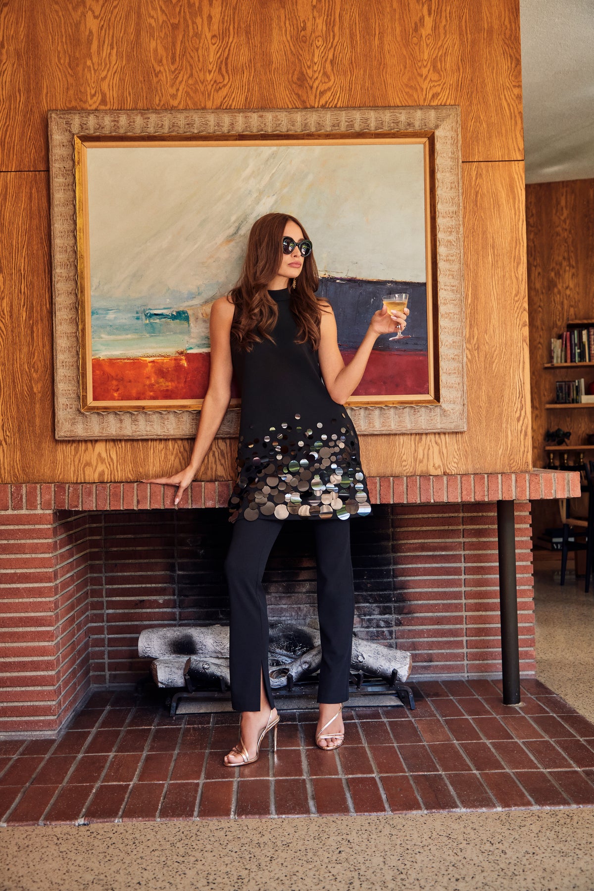 A woman wears an Allena Dress, a sleeveless black tunic with large metallic circular embellishments, styled with sunglasses and black trousers while holding a cocktail glass in front of a brick fireplace.