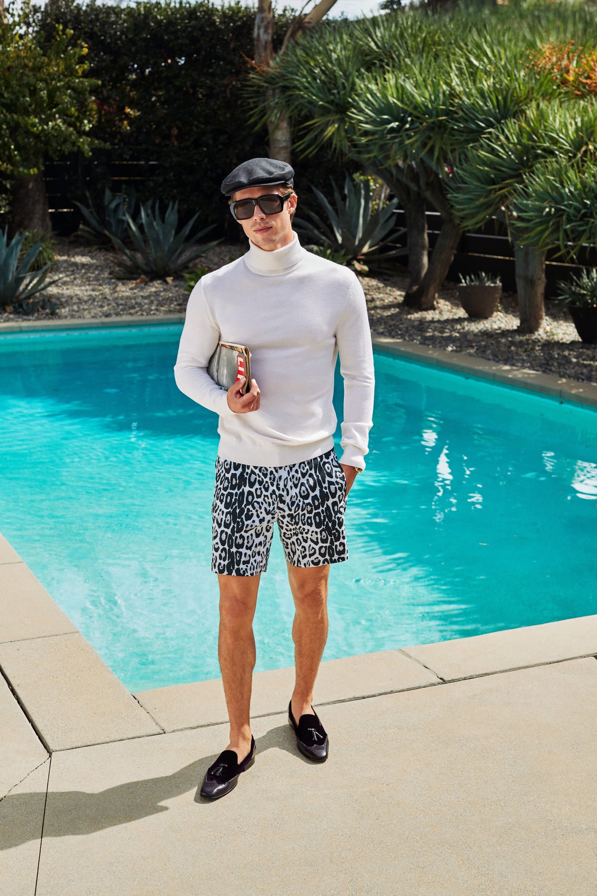 A man wearing a white turtleneck, a flat cap, and grey leopard-print HONOLULU BOARDSHORT stands by a swimming pool holding a clutch.