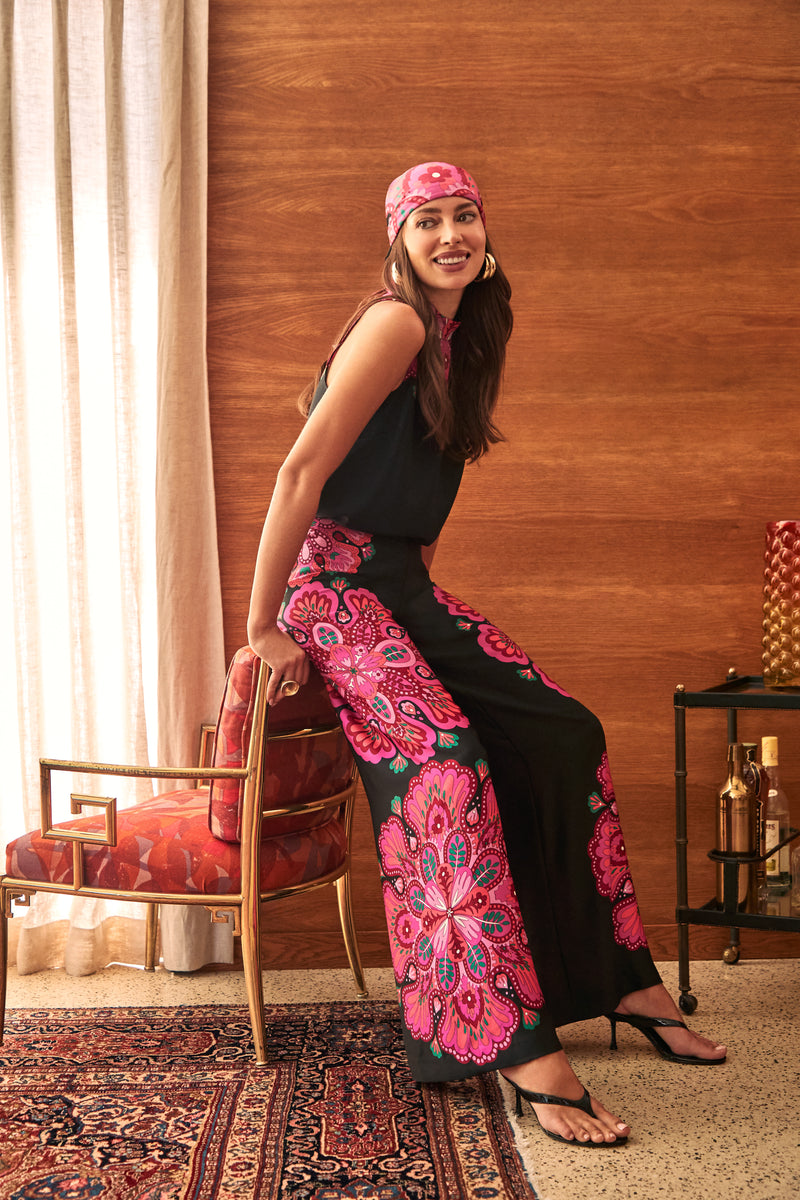 A woman wearing a matching headscarf and Lanay Pant wide-leg trousers featuring a vibrant pink and purple floral pattern, sitting on a patterned chair.