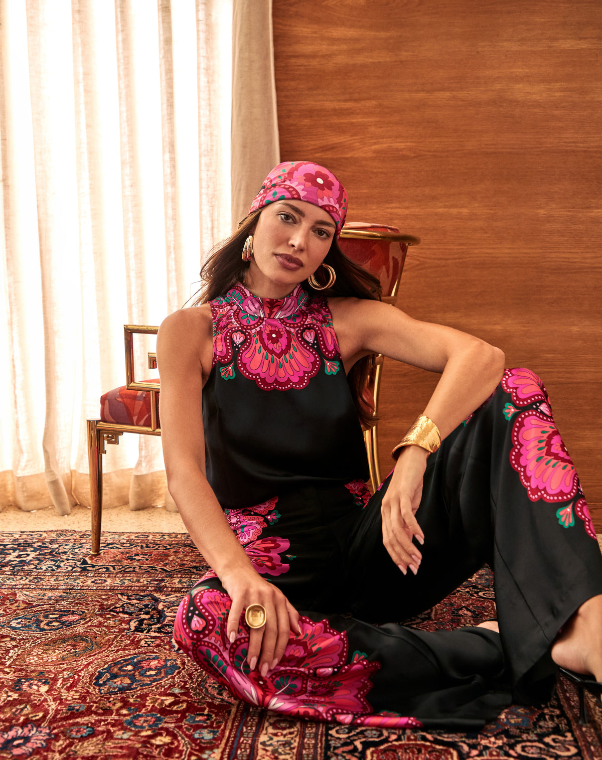 A woman sitting on a patterned rug wears a black sleeveless top and matching wide-leg Lanay Pant featuring a bright pink and orange floral print, paired with a matching headscarf.